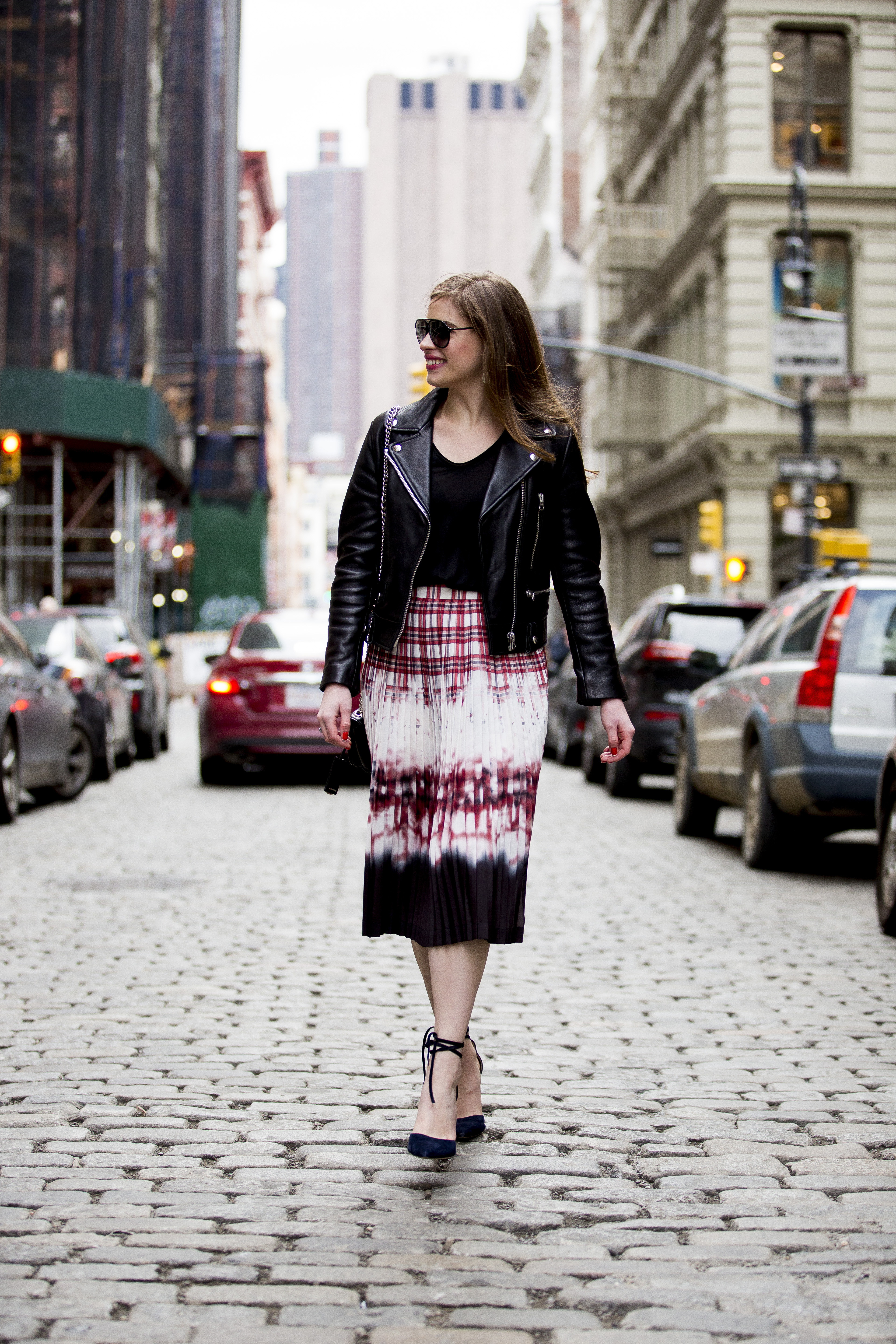 Woman walking down a cobblestone street in Manhattan wearing a black leather jacket, black t-shirt, red, black & white printed skirt, navy heels and black sunglasses. She has brown hair