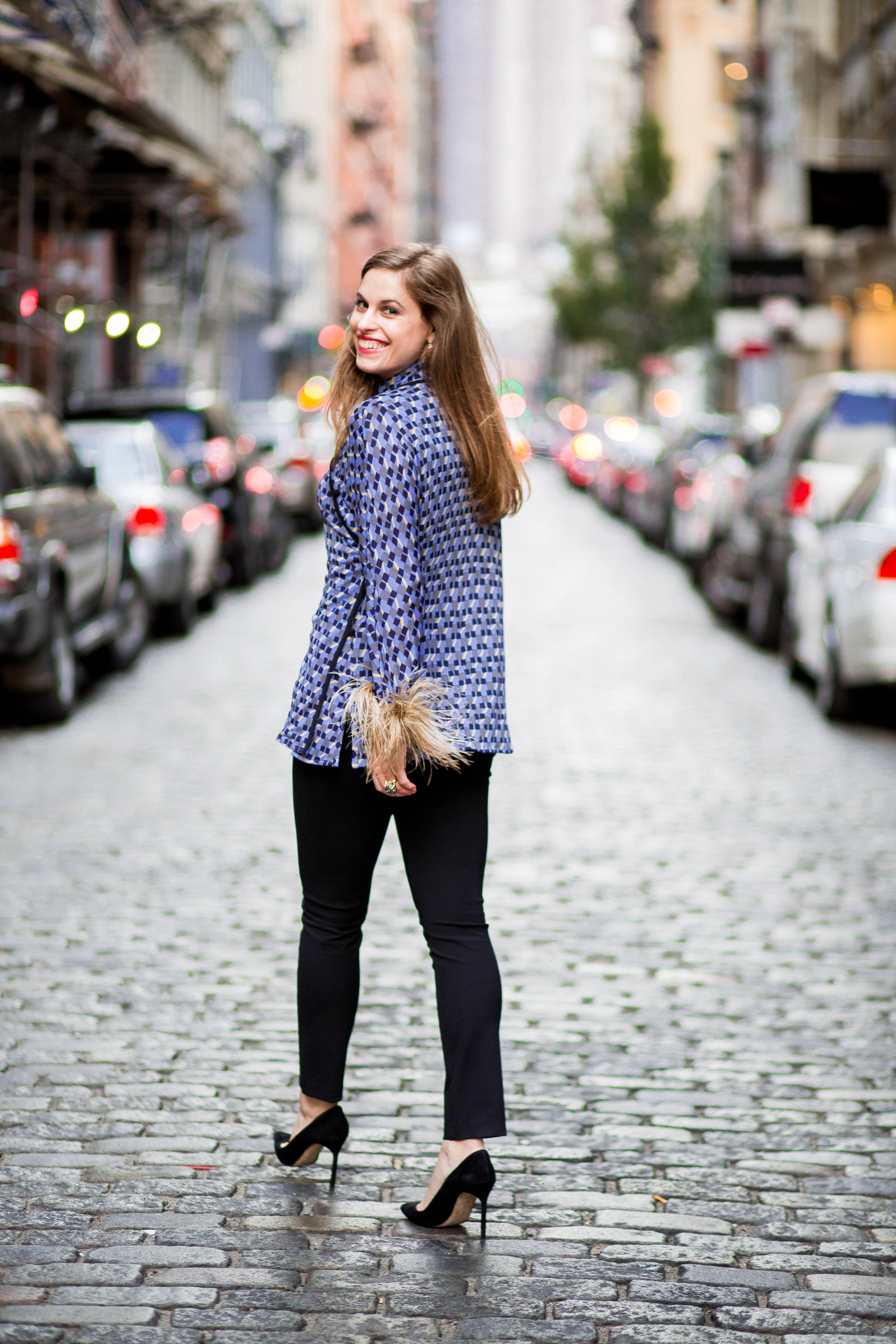 Woman walking down the street in Manhattan away from the camera, but she has turned around: she is smiling, has brown hair, wears a blue, black and white top with feathers on the sleeves, black pants and black pumps