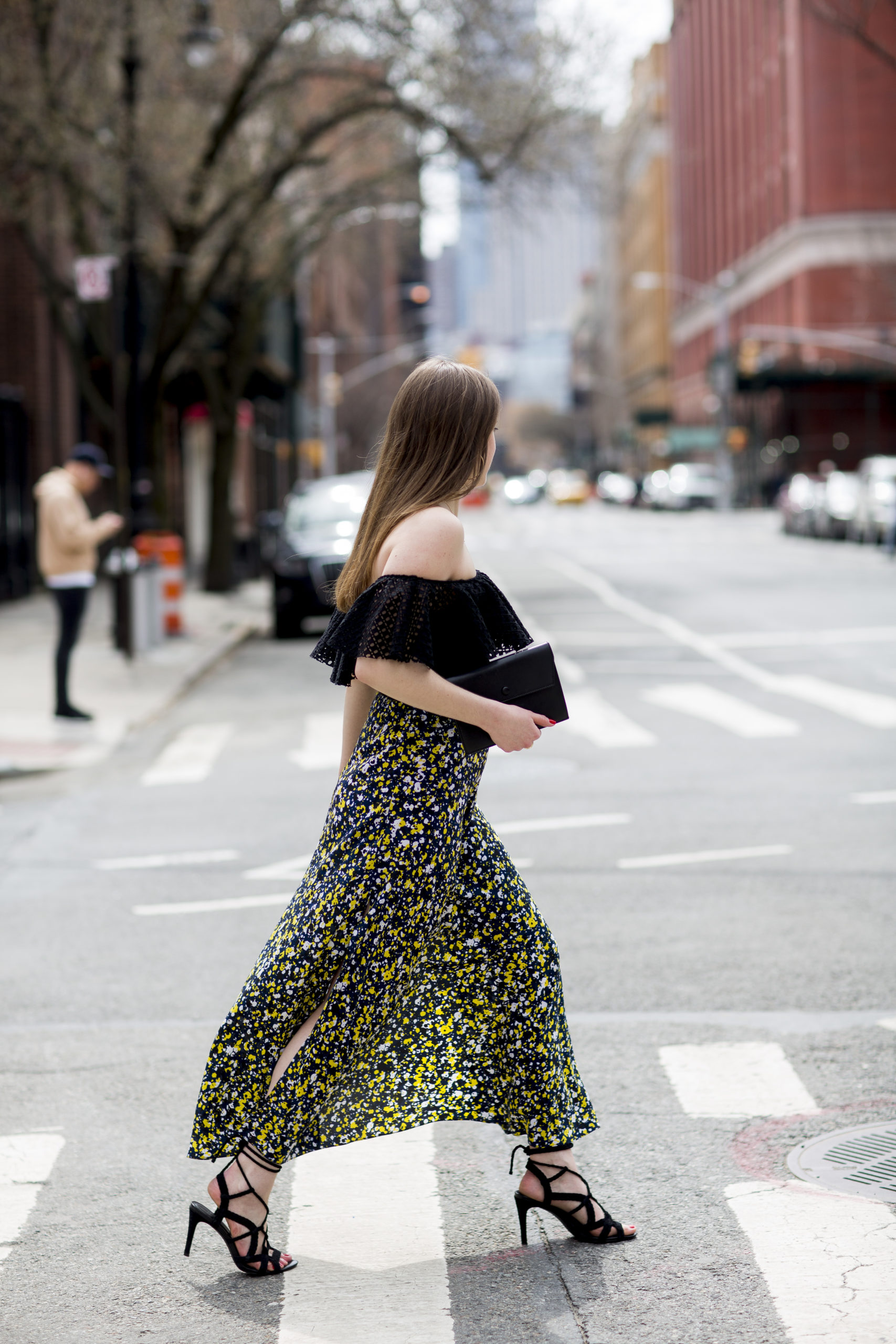 Woman walking across a crosswalk in NYC. She has brown straight hair and is wearing an off-the-shoulder black eyelet top, a printed black, yellow and blue skirt with slits and black heels