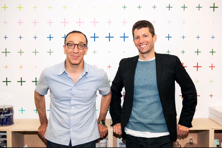 Two men: one wearing glasses, a blue short sleeve button down and jeans, the other wearing a black blazer & a blue t-shirt leaning against a desk with a white backdrop