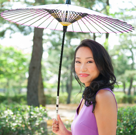 Asian woman standing in a park with trees wearing a purple top, she has black hair and is holding an umbrella