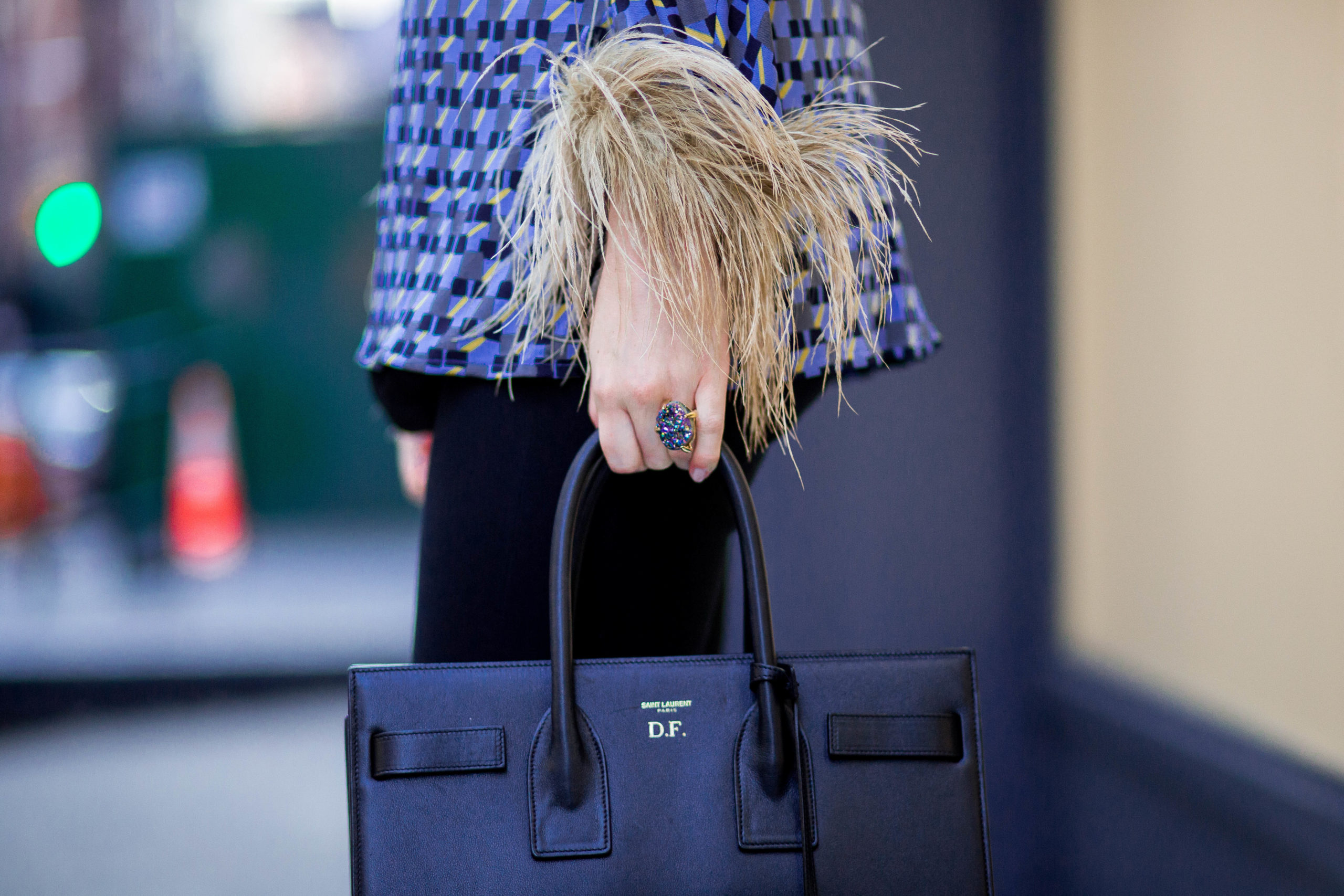 Photo of a woman crossing the street: she is wearing a blue, black & white top with feathers on the sleeve, a large cocktail ring, black pants & a black Yves Saint Laurent bag