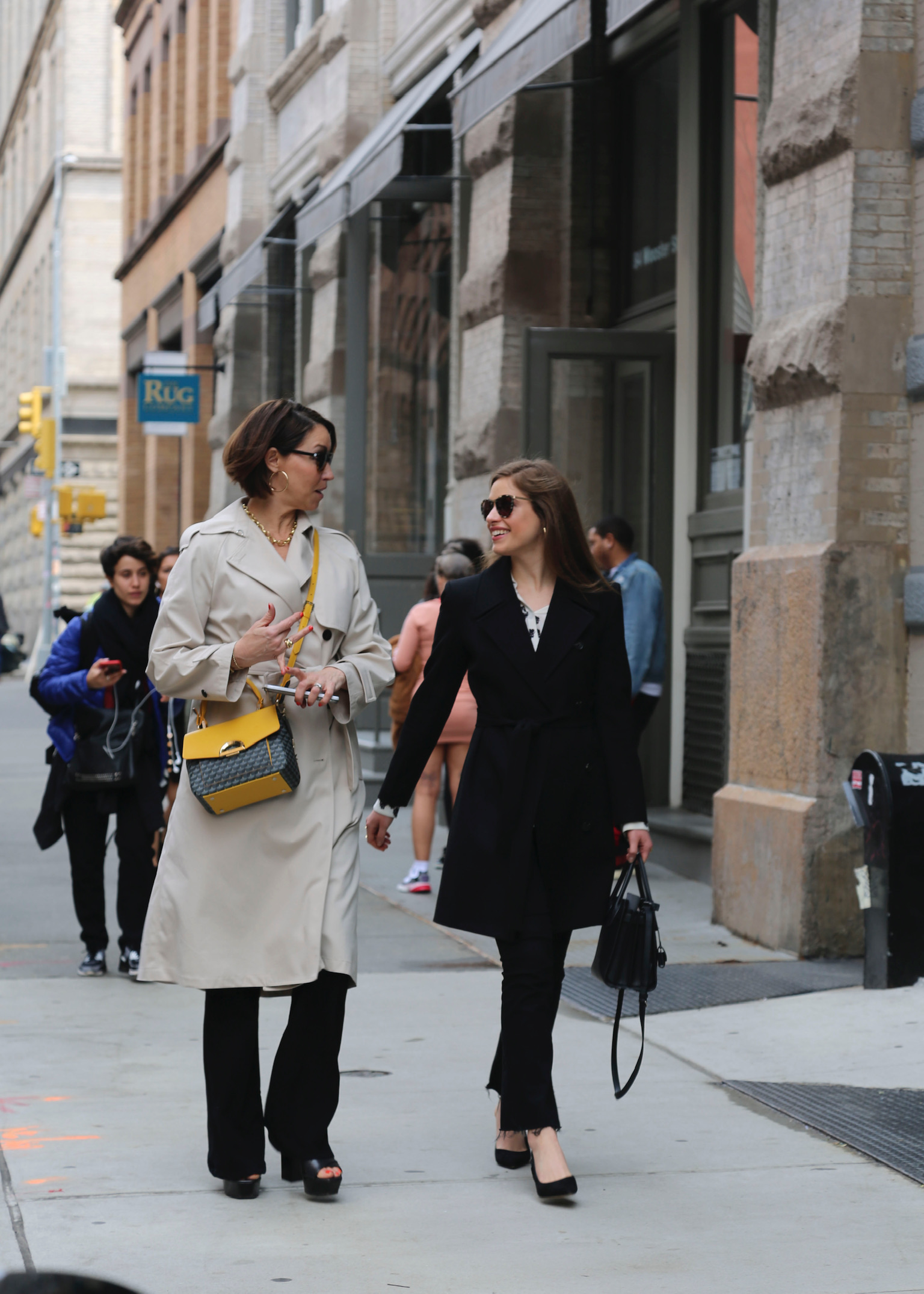 Two women walking down the street in Manhattan: both with brown hair - one wearing a trench coat and has a grey & yellow crossbody bag with black jeans and black wedges; the other wears a black trench coat over a white tea, black jeans and black wedges. They both have on sunglasses