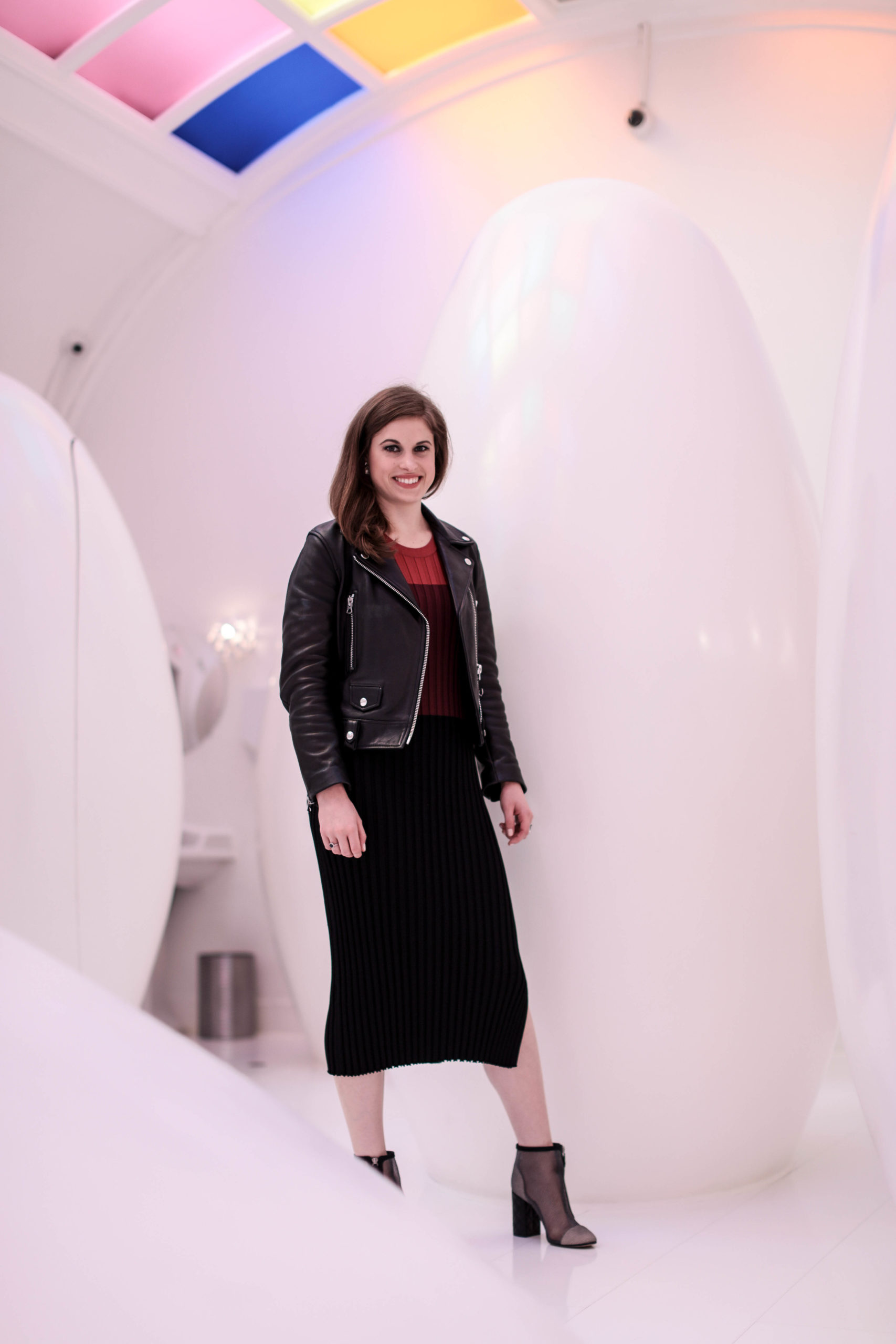 Woman standing in the bathroom at sketch in London in front of the egg-shaped sculptures: she has brown hair, wears a black leather jacket, a black & red striped dress and botties