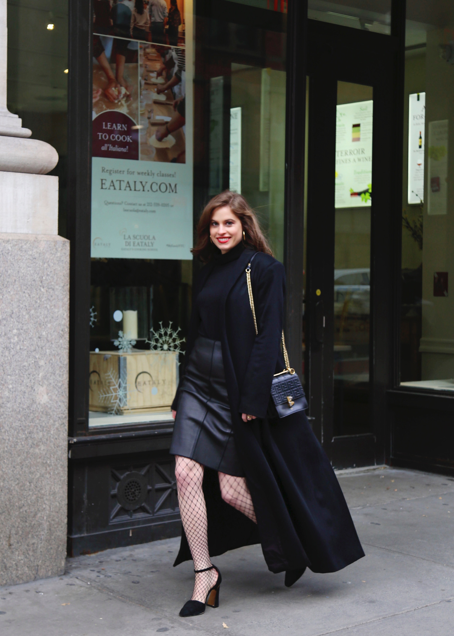 Woman walking down the street in Manhattan: with brown hair wearing a black coat, black turtleneck, black leather pencil skirt, black heels and carrying a black bag