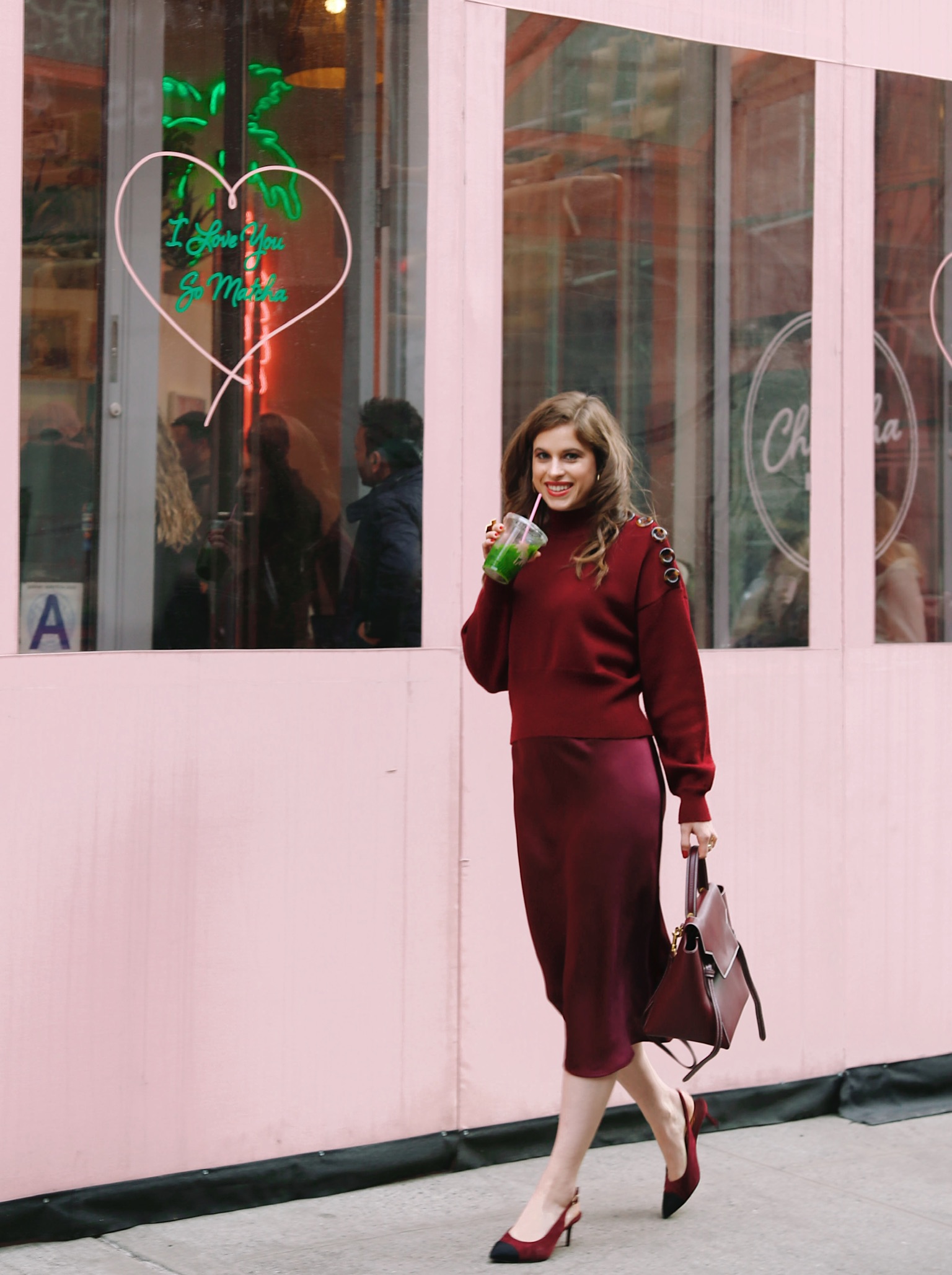 Woman in front of Cha Cha Matcha drinking a matcha drink wearing a crimson monochrome outfit: sweater, handbag, shoes & skirt