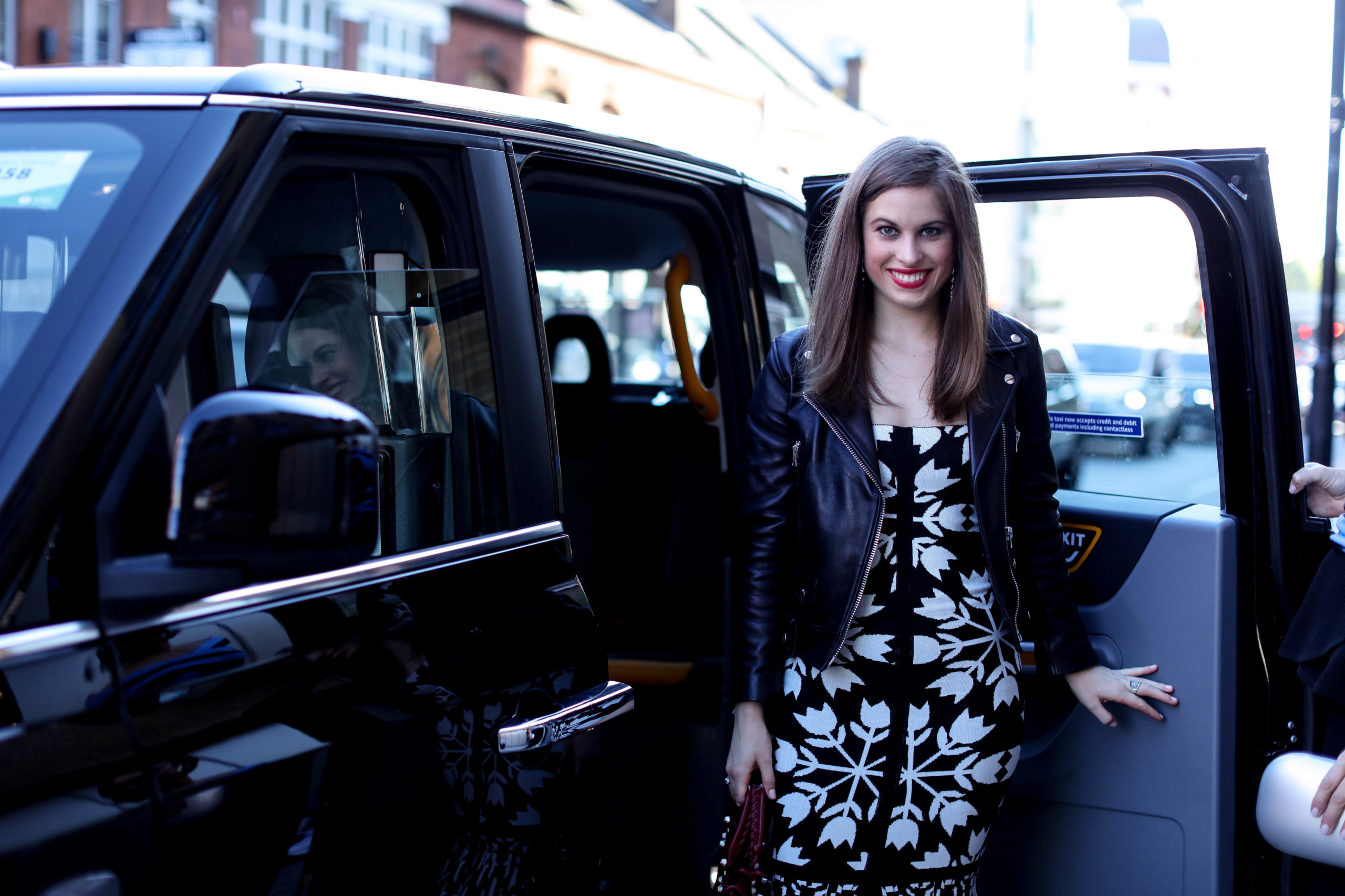 Woman with brown hair getting out of a taxi cab in London wearing a black leather jacket and a black & white printed dress