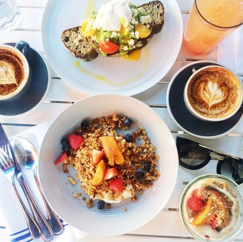 Delicious looking brunch spread at Bluestone Lane: avocado toast, two lattes with heart latte art and a french toast on a white wooden picnic table with sunglasses on the table