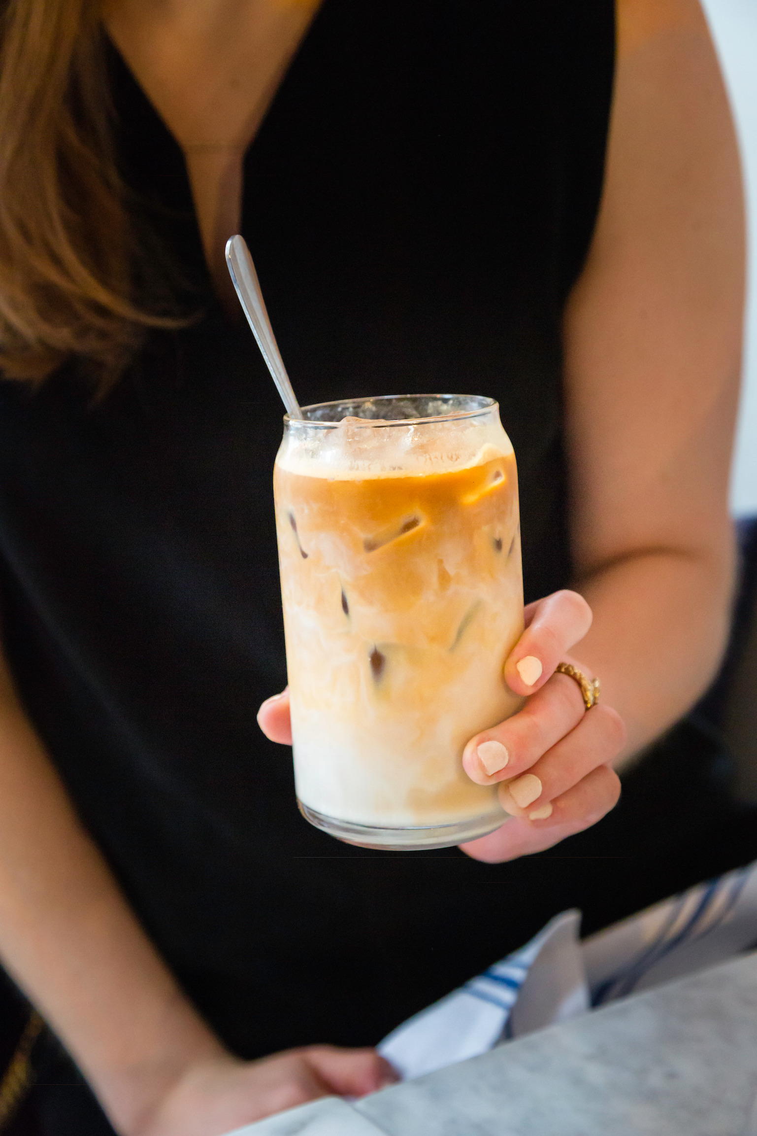 Woman sitting at a table wearing a black sleeveless dress, holding an iced coffee
