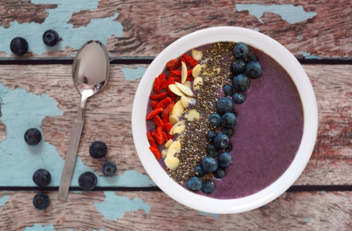 açaí bowl with goji berries, almond slivers, chia seeds, blueberries with a spoon on a wooden table with blueberries on the table