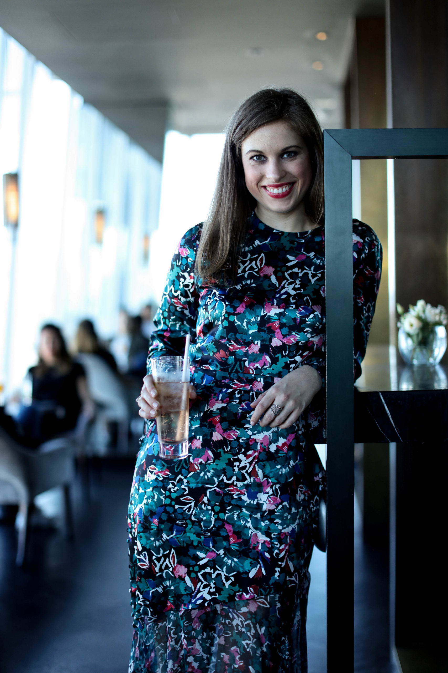 Woman standing at a bar: she has brown straight hair and is wearing a blue & pink printed dress. She is holding a drink