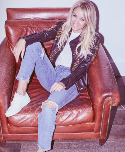 Woman sitting in a chair wearing a black leather jacket over a white t-shirt, jeans and white sneakers. She has blonde curly hair and is smiling