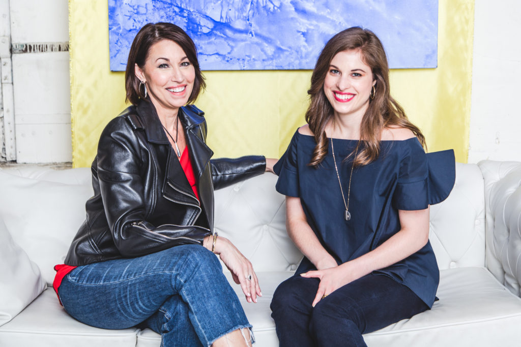 Two women sitting on a white couch with artwork: one wears a black leather jacket with a red top underneath and jeans, the other wears an off-the shoulder navy top, a pendant necklace and jeans they are smiling and both have brown hair