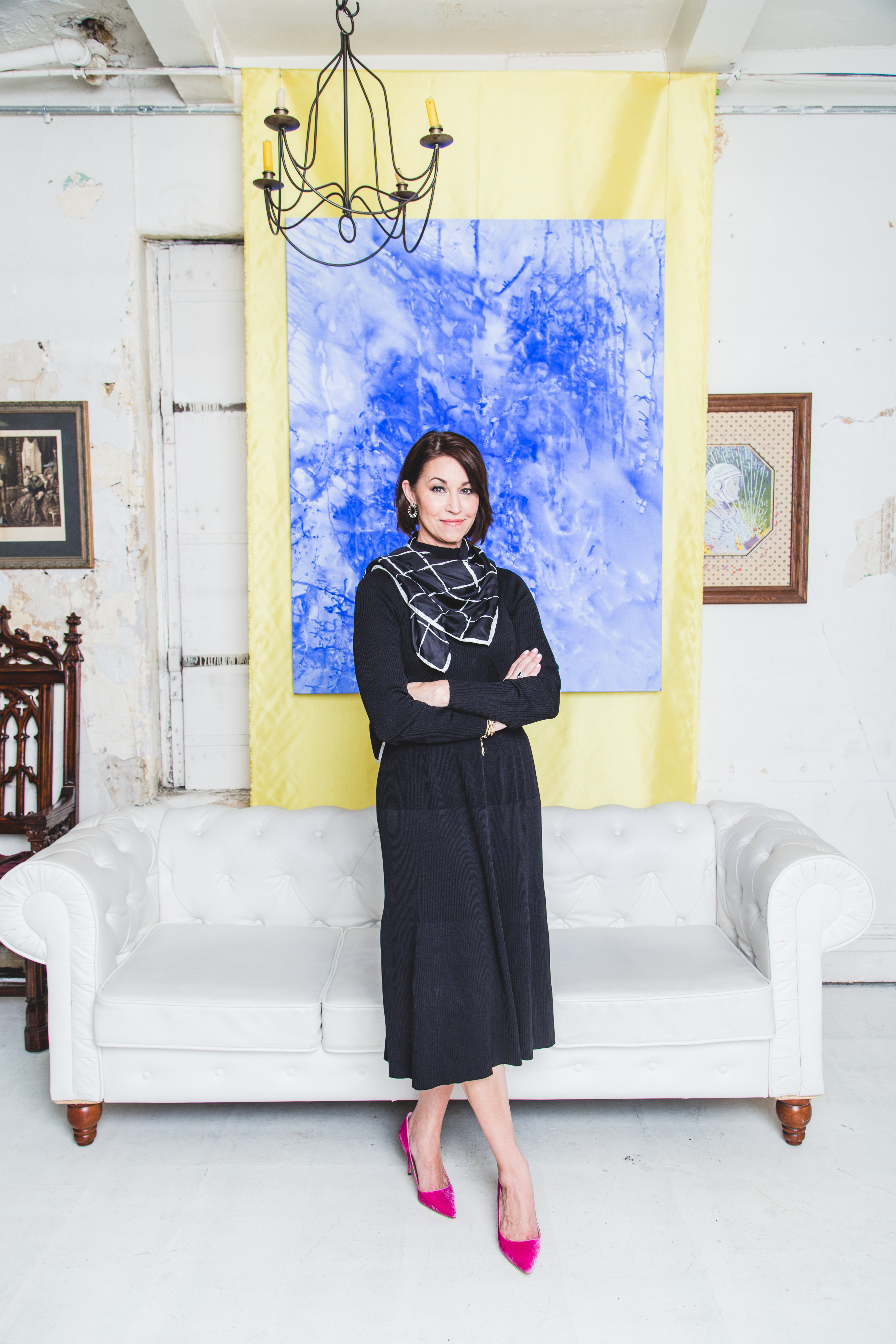 Woman standing in front of a white couch with artwork on the wall: wearing a black dress, printed scarf and pink pumps