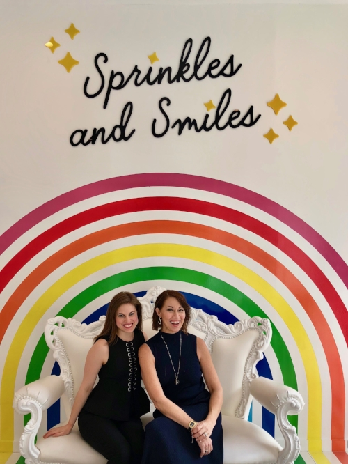 Two women sitting on a white couch both wearing all black in front of a rainbow that says: sprinkles & smiles
