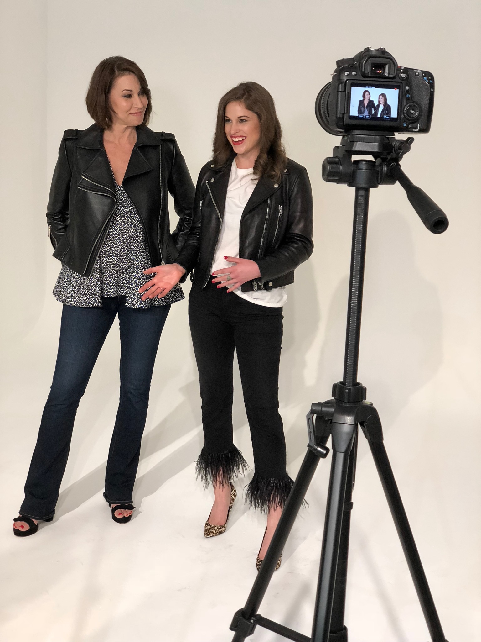 two women on set both wearing leather jackets: one has a blue and white printed top underneath, the other has a white t-shirt, jeans with feathers and burberry pumps