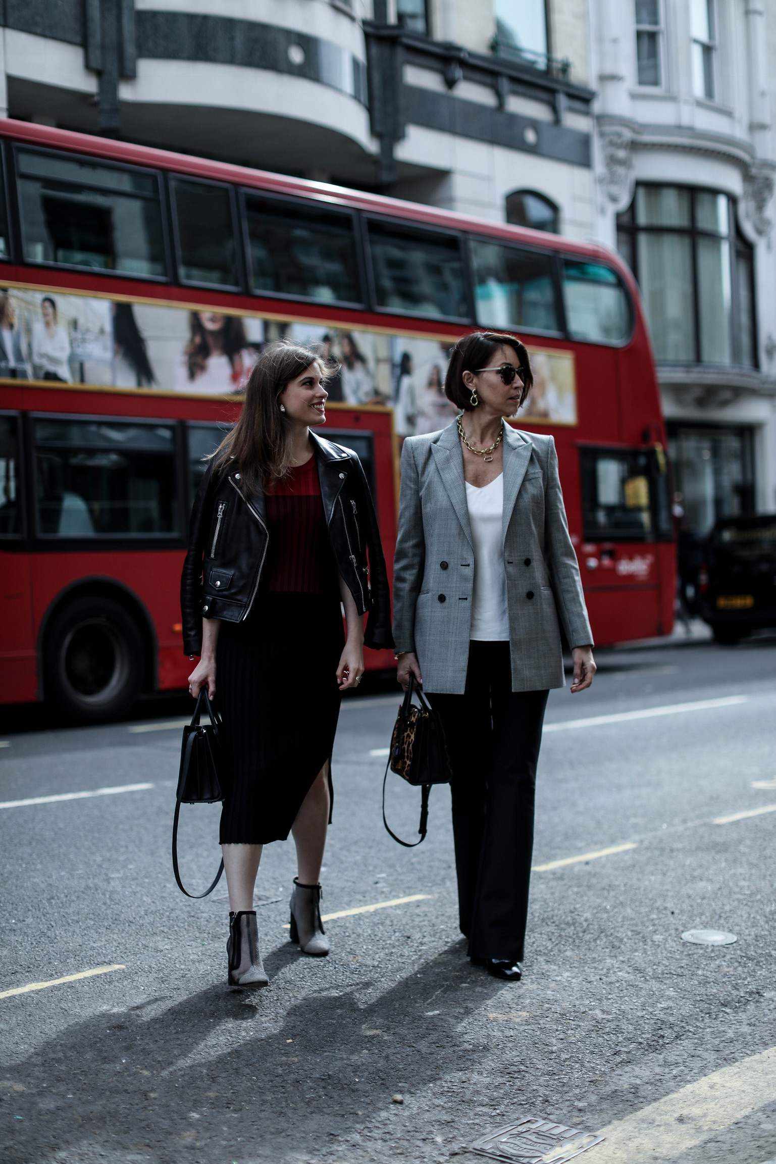 Two women walking in front of a red double decker bus in London: one wearing a black leather jacket, red & black stripe dress and booties, the other wears a plaid blazer with a white cami and black pants. They both have brown hair, are wearing sunglasses and carrying square bags