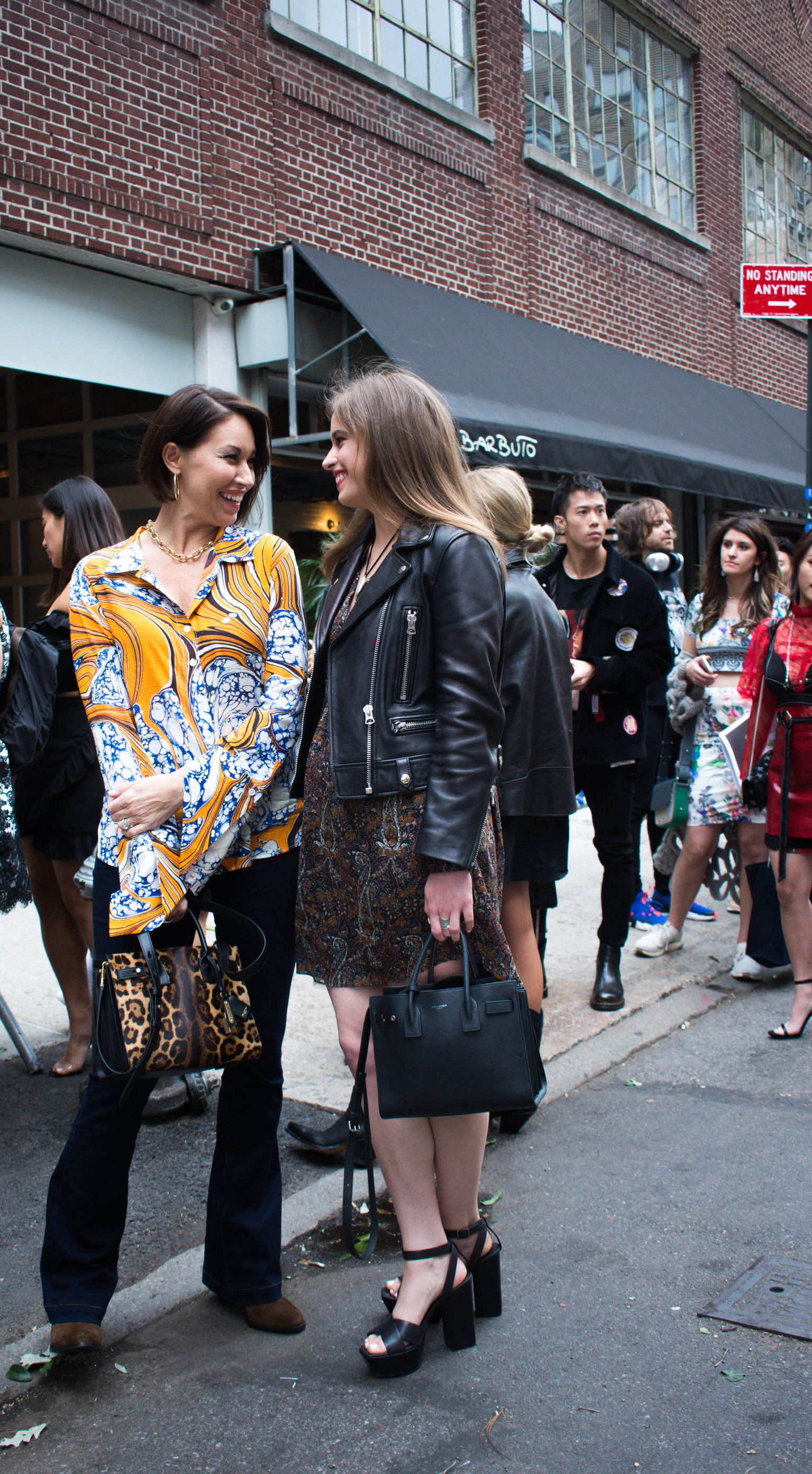 Two women standing outside a fashion show: one wearing an orange, white and blue button down top and the other is wearing a black leather jacket, printed dress and wedges