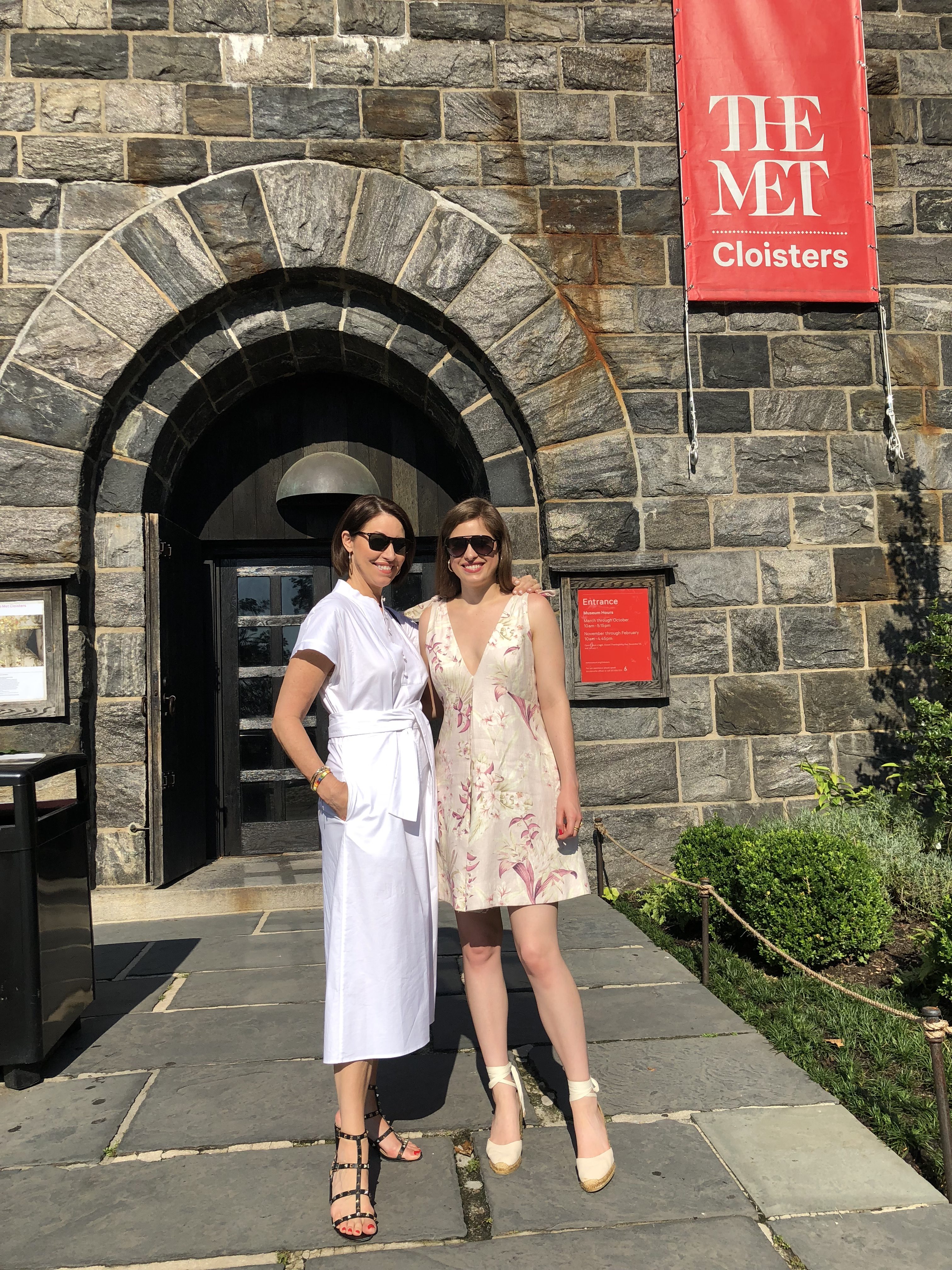 Two women standing in front of The Cloisters: a woman in a white dress and a woman in a v-neck printed dress, both in wedges and sunglasses and both have brown hair
