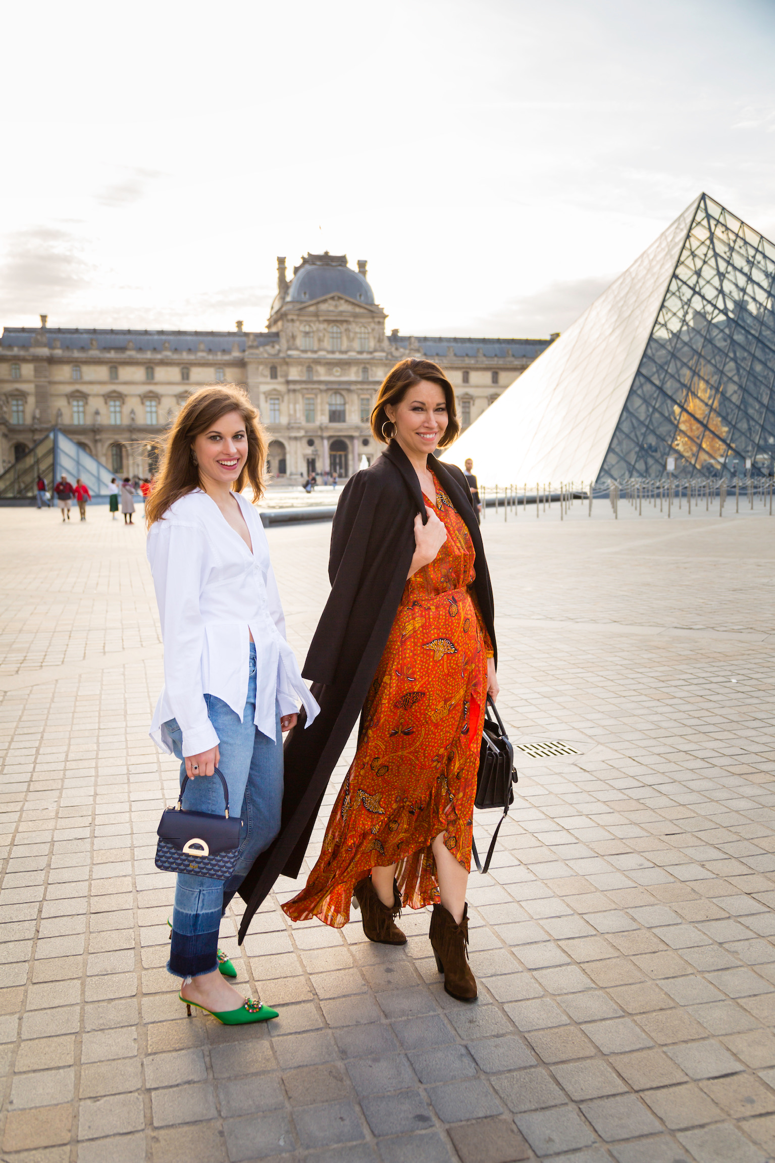 Two women walking by The Louvre: one wearing a white top, jeans and green kitten heels, the other is wearing a black blazer and a printed dress with brown cowboy boots