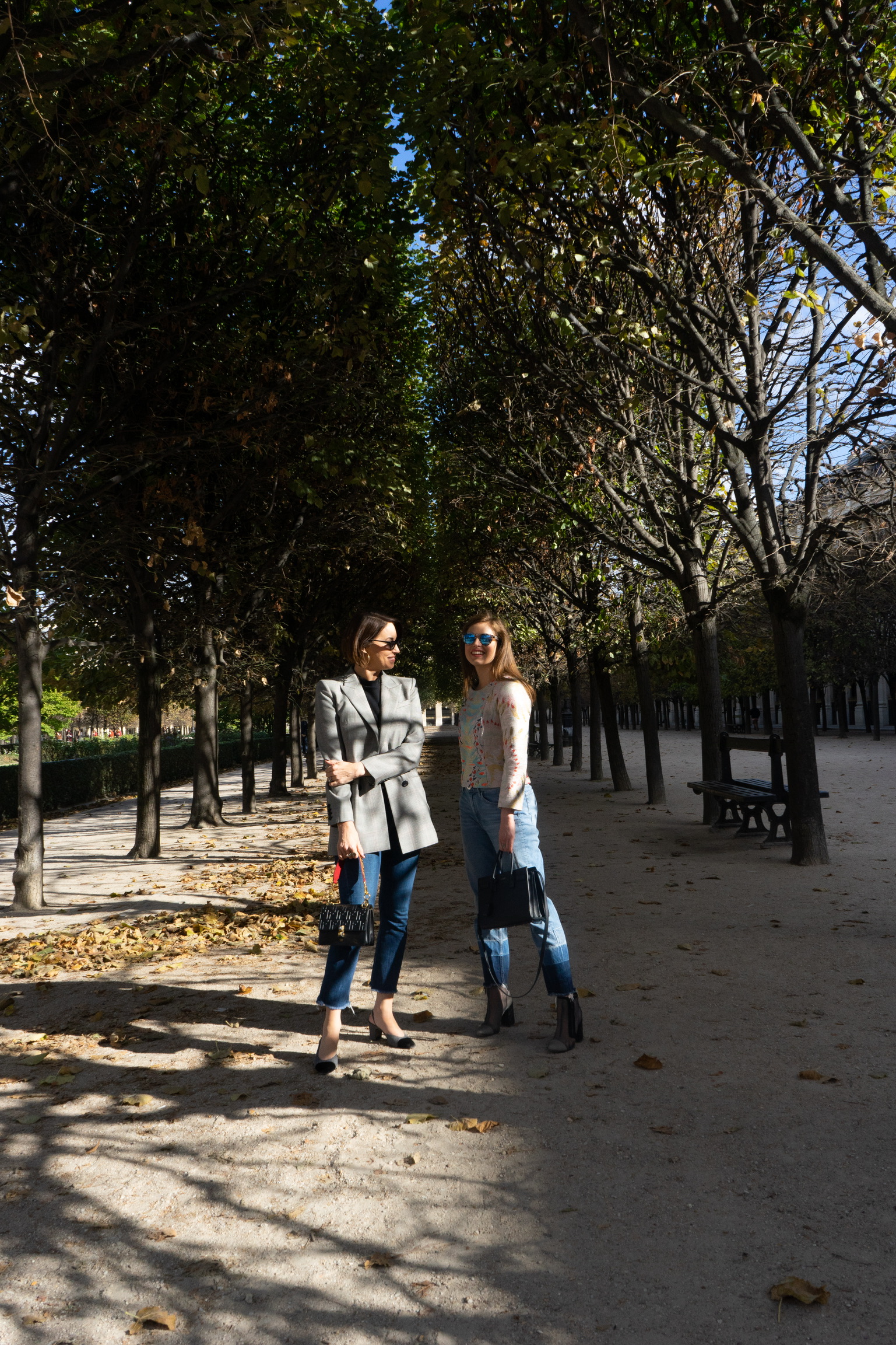 Two women standing in a park: one wearing a blazer and jeans and the other wearing a printed top and jeans