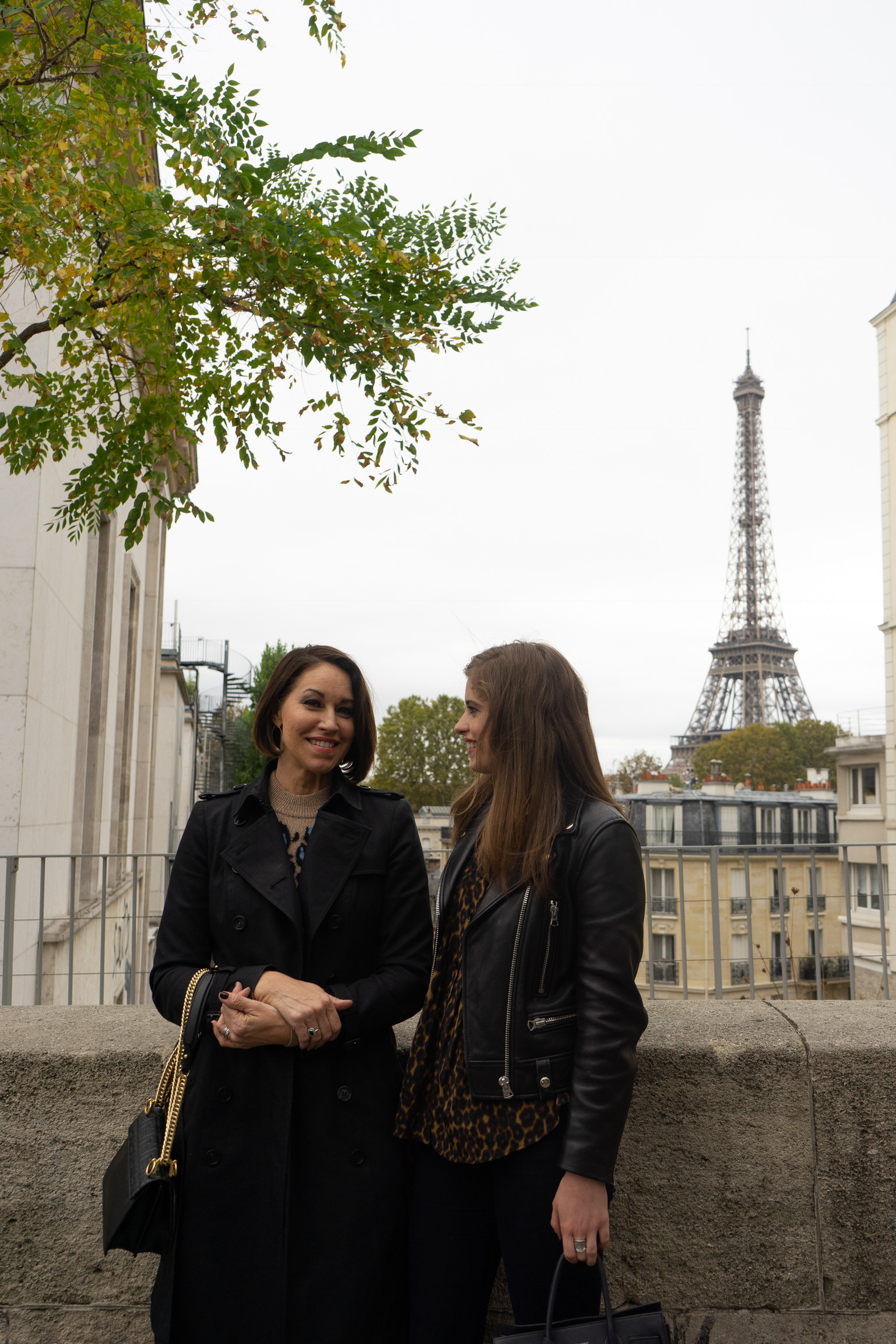 Two women standing in front of the Eiffel Tower: one wears a black trench holding a black purse, the other wears a black leather jacket, leopard print top and black jeans. They both have brown hair