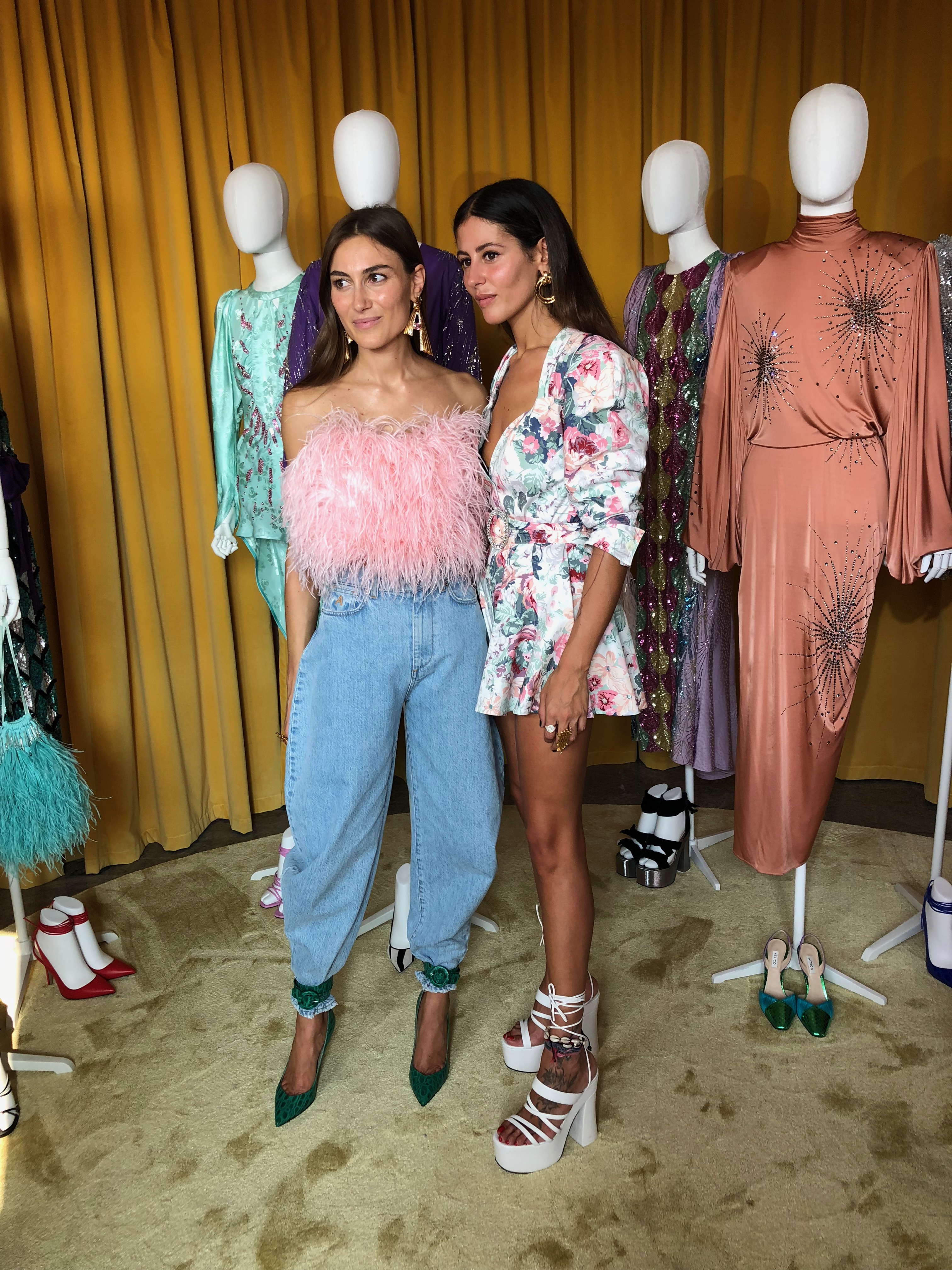 Two women standing in front of mannequins: one wearing a pink feather top with jeans and green heels, the other wearing a mini floral print dress and white wedges