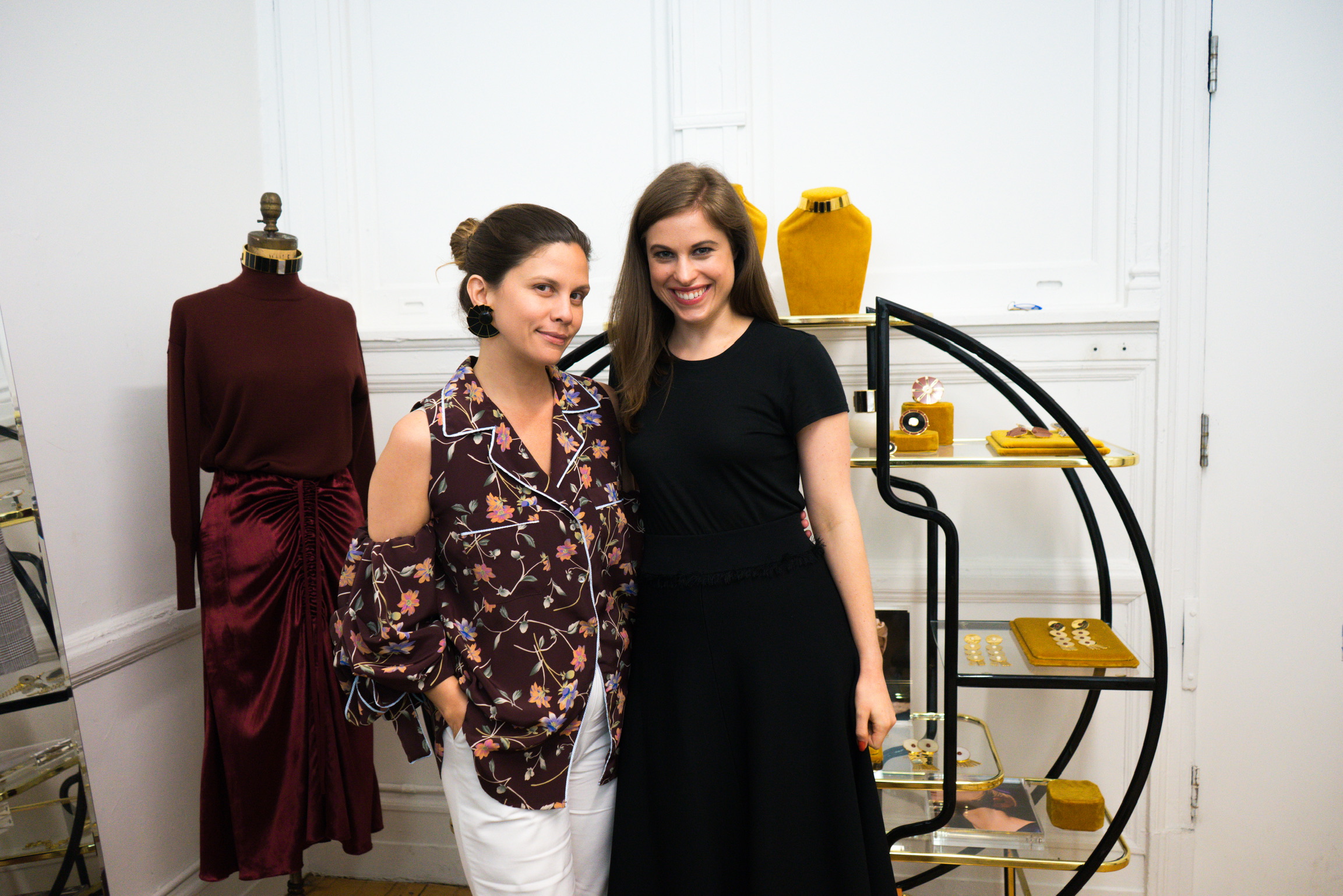 Two women standing in front of a jewelry display & a mannequin: one wearing a printed top & white jeans, the other wearing an all-black look - they both have brown hair