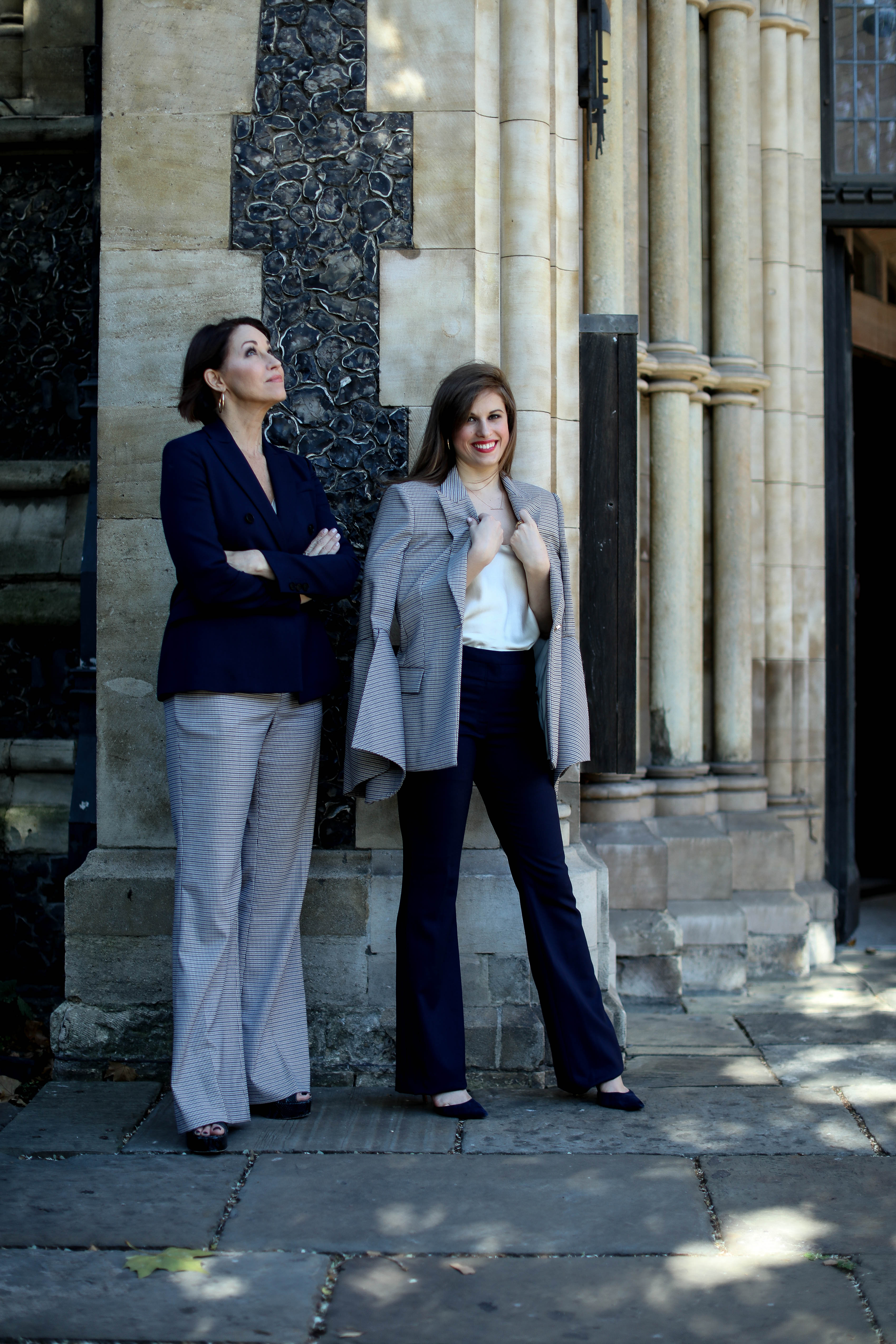 Two women standing in front of a church in london: one wearing a navy blazer & plaid pants, the other wearing a plaid blazer with a white cami and navy pants. Both in high heels and have brown hair