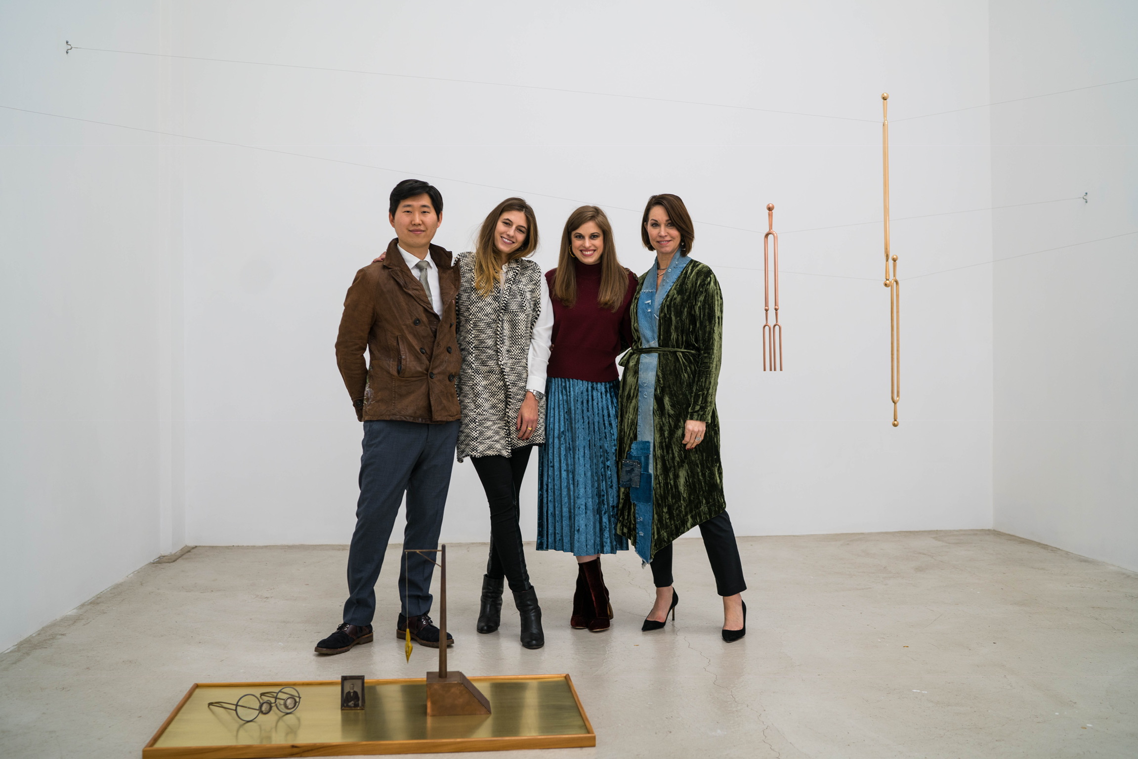 Four people (one man and three women) standing in an art gallery: one wearing a brown jacket, blue pants and brown shoes, one wearing a scarf and jeans, one wearing a crimson sweater and blue skirt, the last wearing a green velvet robe coat with denim on it and black pumps.