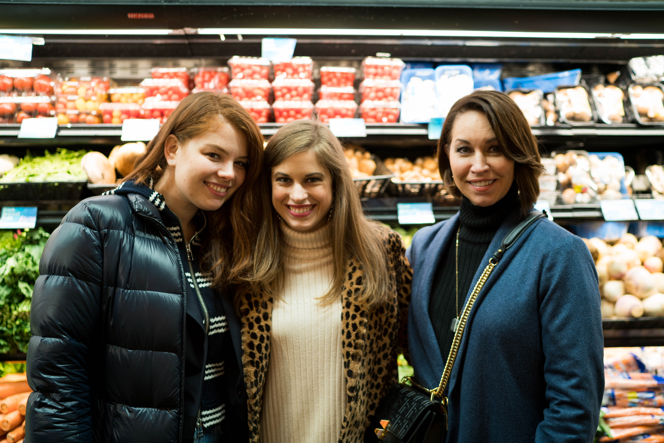 Three women in a grocery store: one wearing a puffer jacket, one wearing a cream turtleneck sweater and leopard print coat, the last is wearing a blue coat and a black turtleneck sweater. One has red hair and the other two have brown hair
