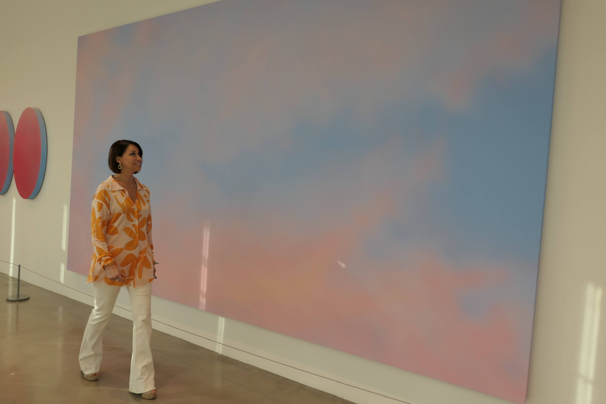 Woman wearing an orange and pink long sleeve printed top, white wide leg jeans and wedges walking past artwork