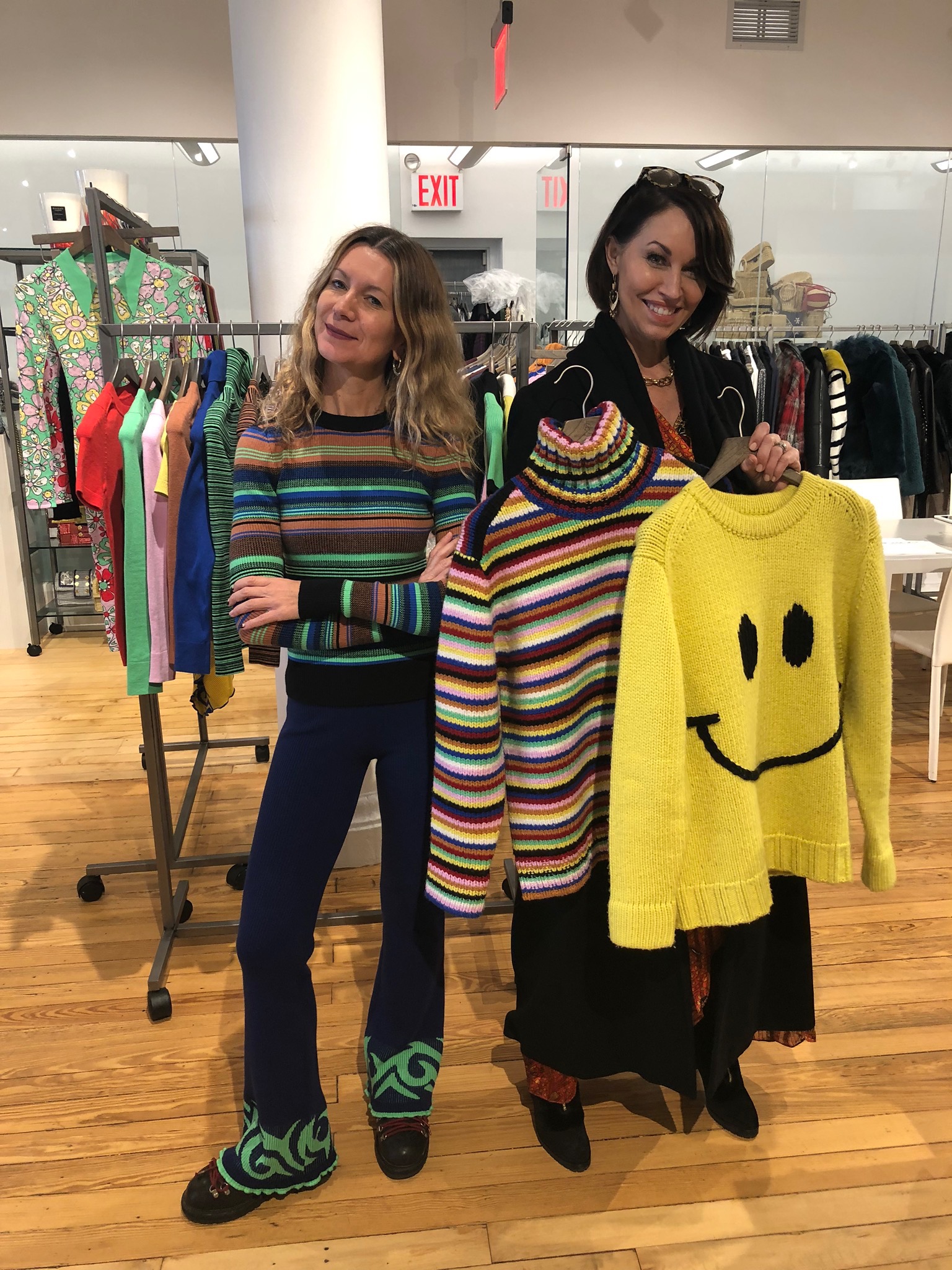Two women: one wearing a rainbow stripe sweater, the other is holding up a rainbow strip turtleneck sweater and a yellow smiley face sweater. One has blonde wave hair and the other has short brown straight hair