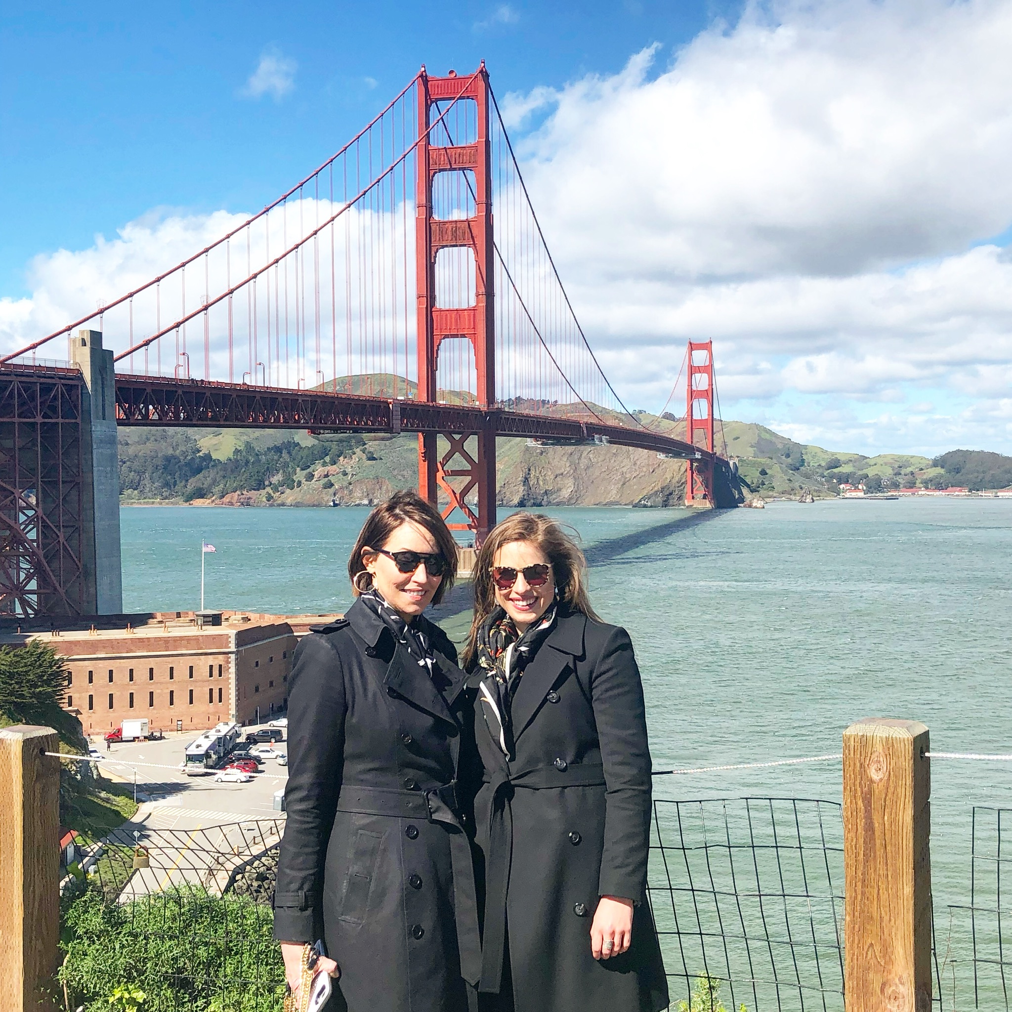 Two women standing in front of the golden gate bridge both wearing sunglasses and black trench coats and have brown hair