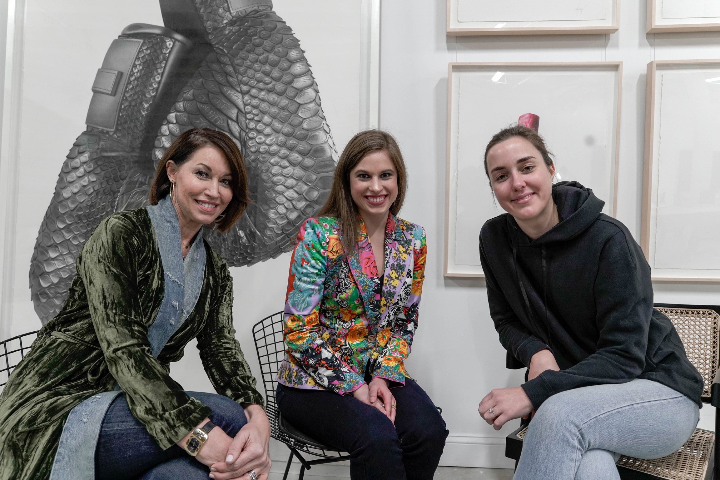 Three women in front of a piece of art: one wears a green velvet robe with denim, another wears a bright printed blazer and black jeans and the other wears a black sweatshirt with blue jeans
