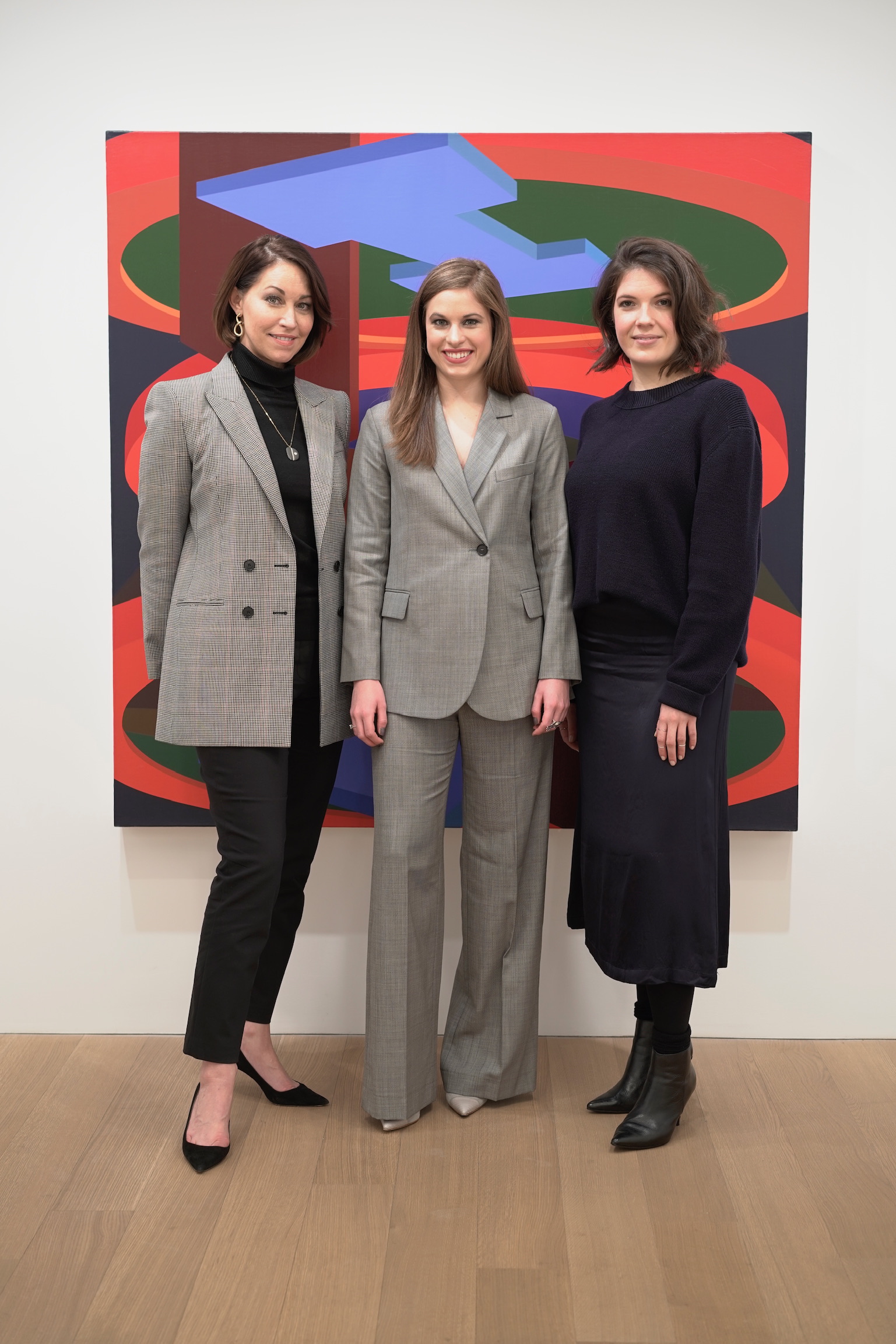 three women standing in front of a painting: one wearing a plaid blazer with black pants, a black top and black pumps wearing a long pendant necklace; the other is wearing a grey suit with grey heels and the last is wearing a navy sweater and skirt with booties. They all have brown hair