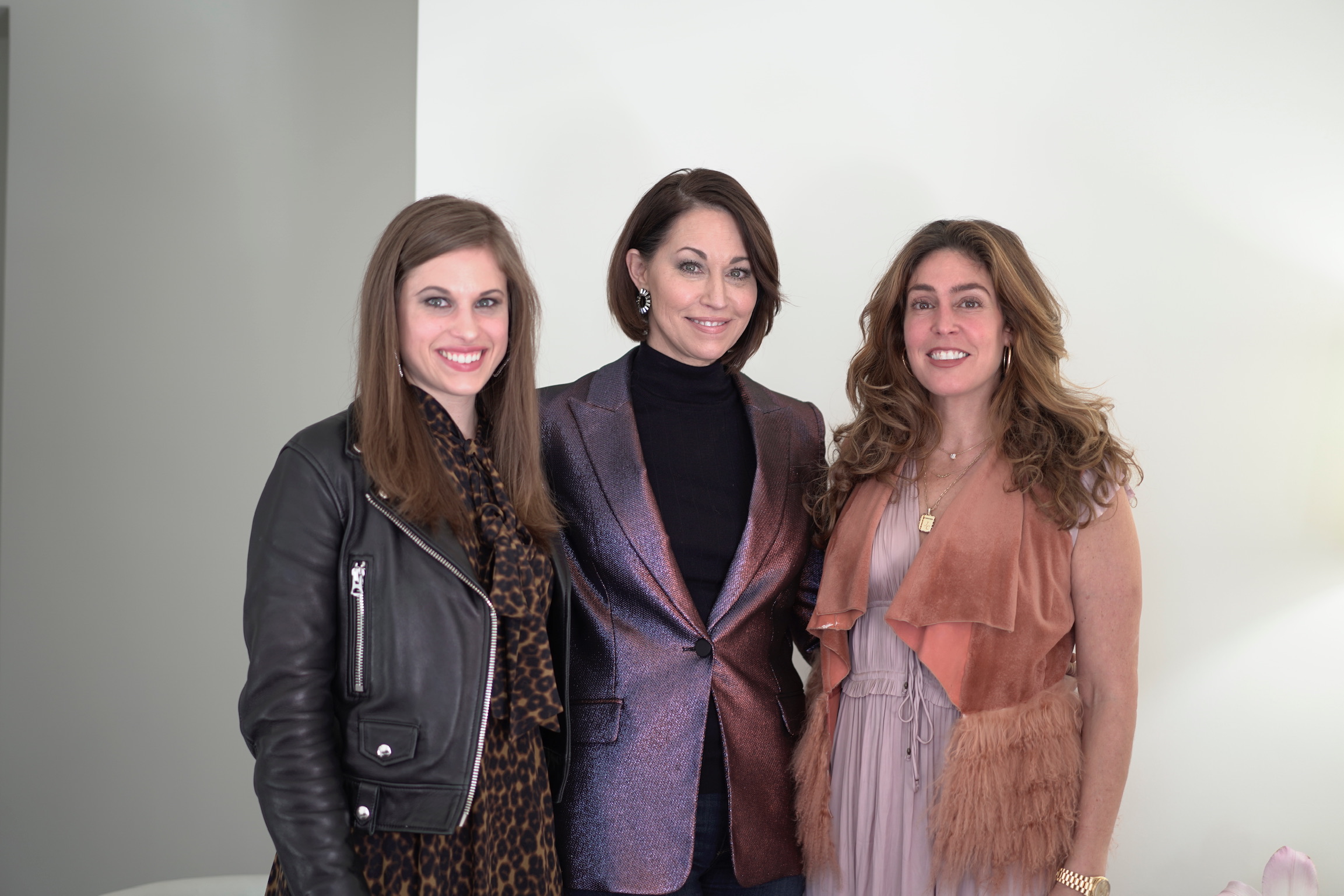 Three women: one wearing a black leather jacket & leopard print dress, one wearing a purple blazer with a black turtleneck and one wearing a pink sleeveless vest over a pink dress. They all have brown hair
