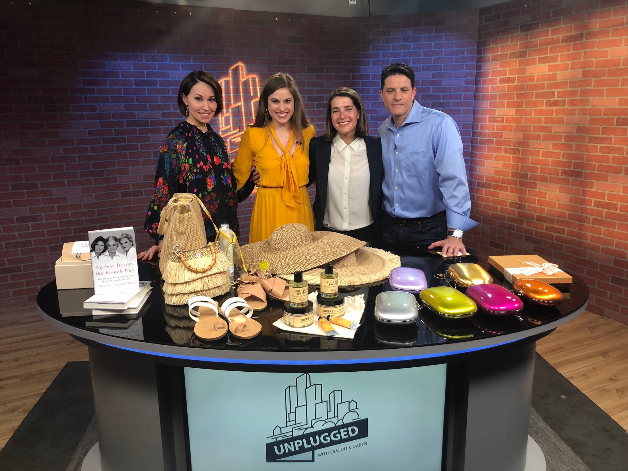 Four people standing on set of a tv station standing behind a counter filled with products: one woman wears a printed top, the other wears a yellow dress, one woman wears a black blazer with a white button down and the male wears a blue button down