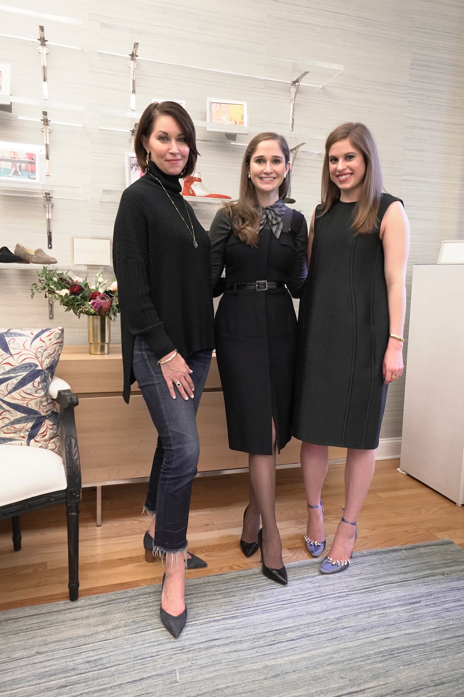 Three women standing in a showroom: one wearing a black top, jeans and heels, and the other two are wearing black dresses and heels, they all have brown hair