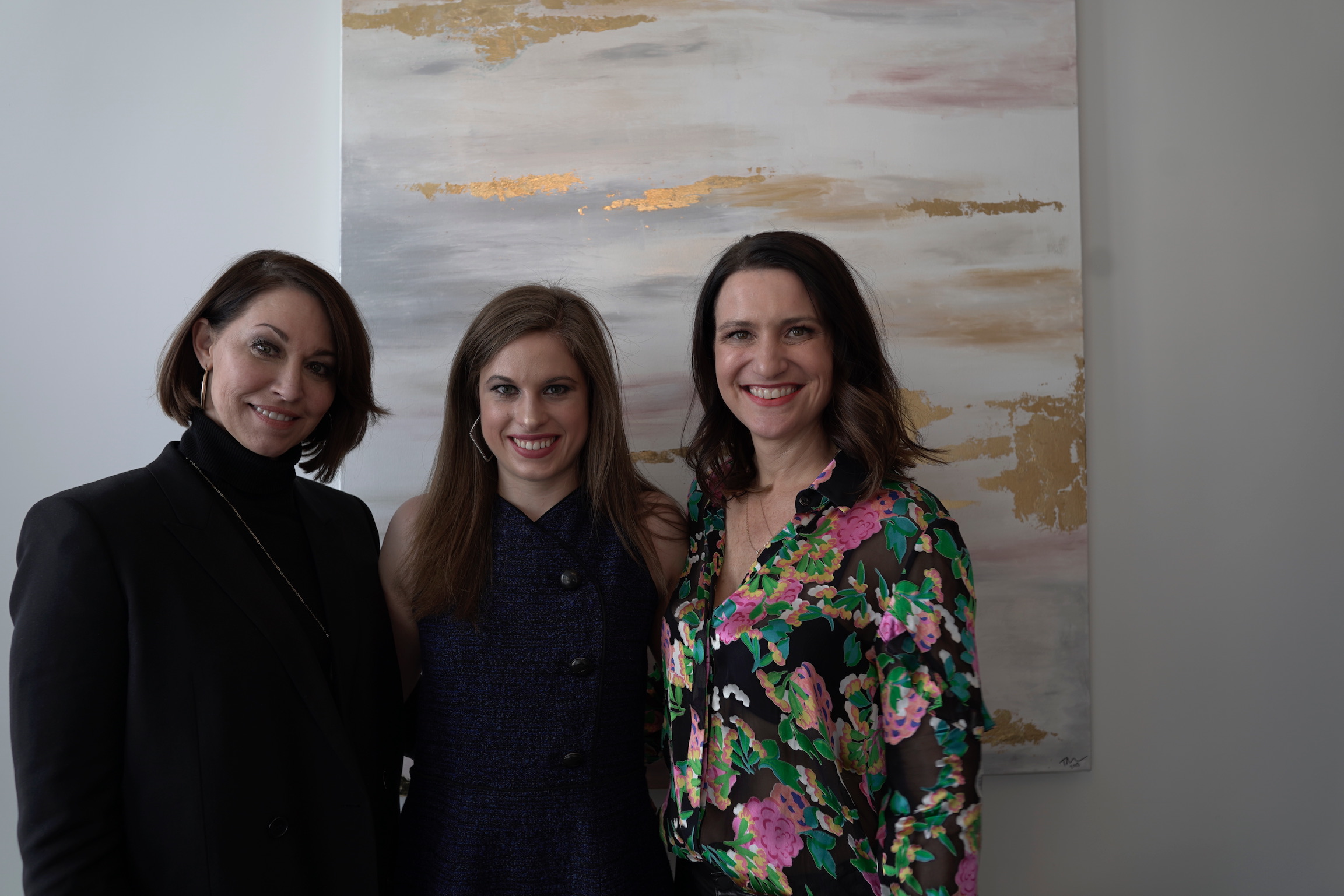 three women standing in front of a painting: one wearing a black blazer, one wearing a sleeveless blue dress and one wearing a printed silk top. All with brown hair