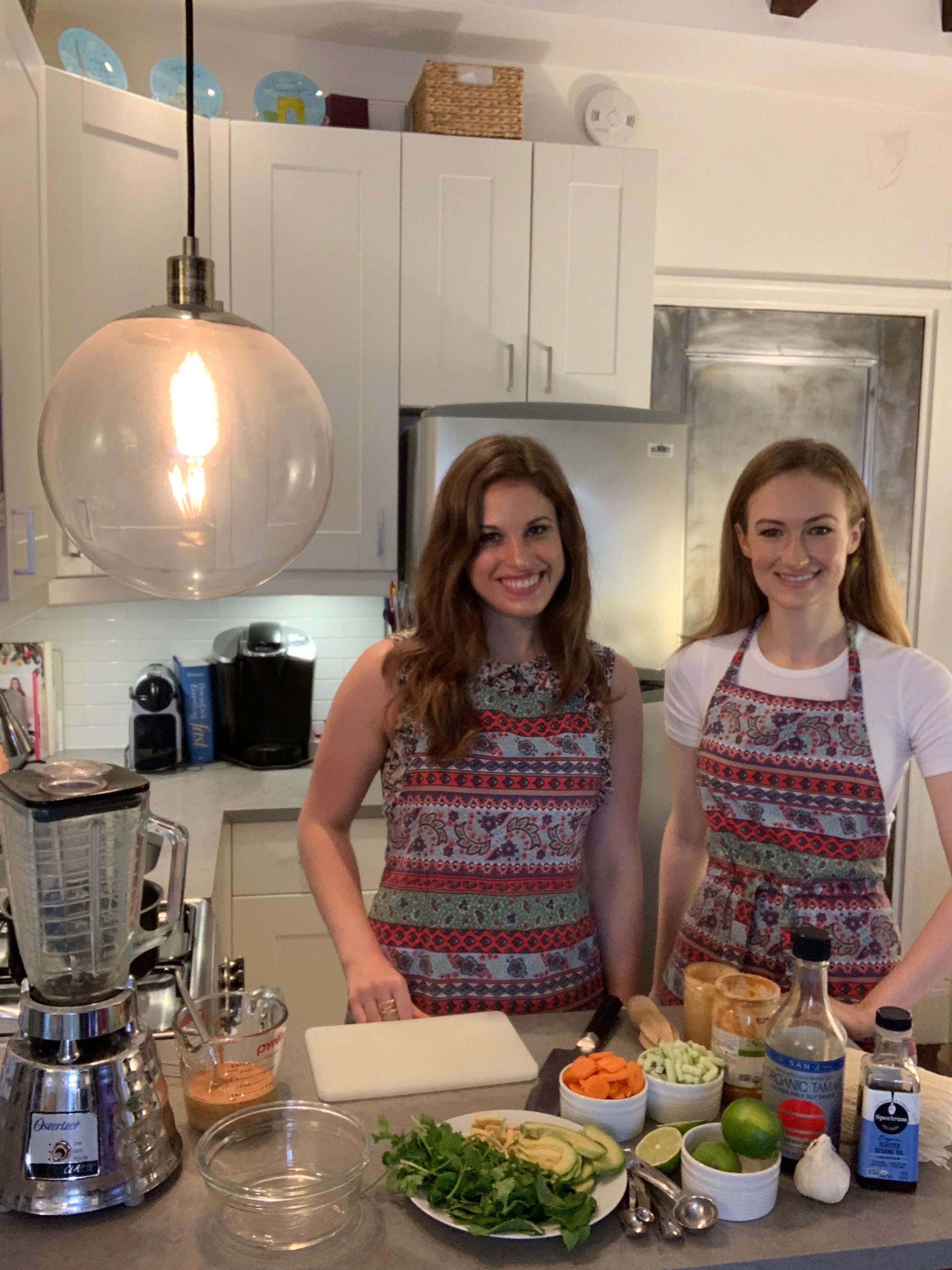Two girls in a kitchen wearing printed aprons. One has curly brown hair and the other has blonde hair