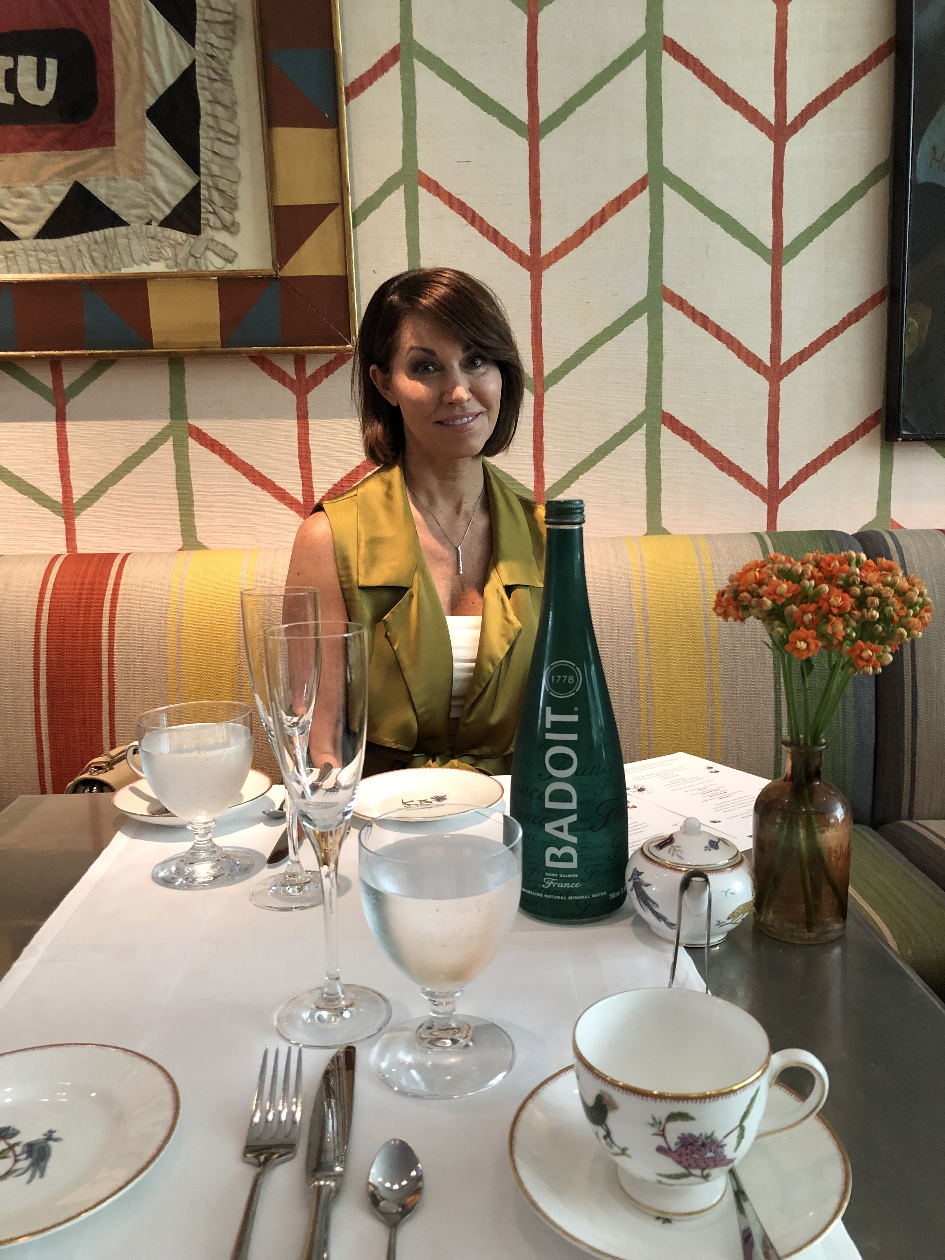Woman wearing a yellow silk sleeveless trench coat sitting at a restaurant table with badoit sparkling water