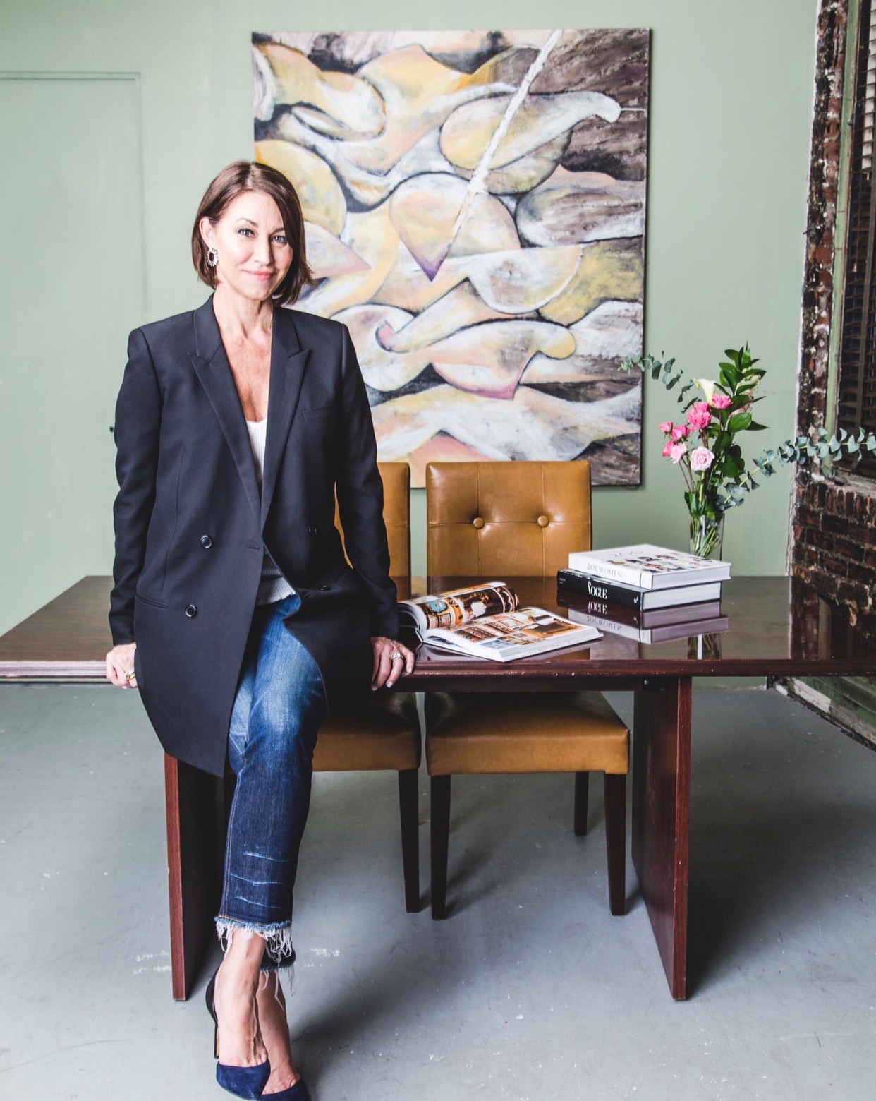 Woman wearing a black blazer & jeans and black pumps sitting on a desk in front of a piece of art