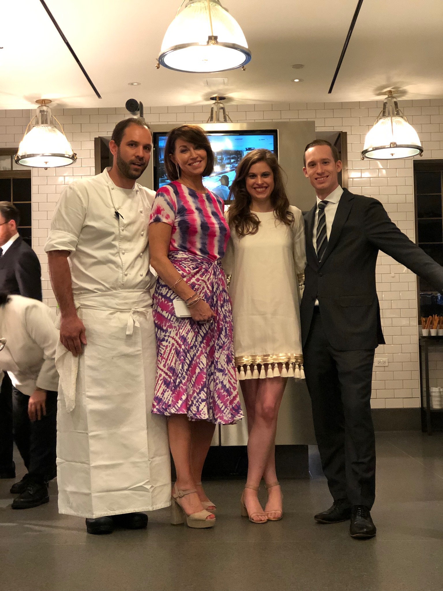 Four people: a man who is a chef in his chef outfit, a woman wearing a tie-dye pink, purple and white outfit, a woman in a cream dress in heels and a man in a suit standing in a kitchen. All have brown hair