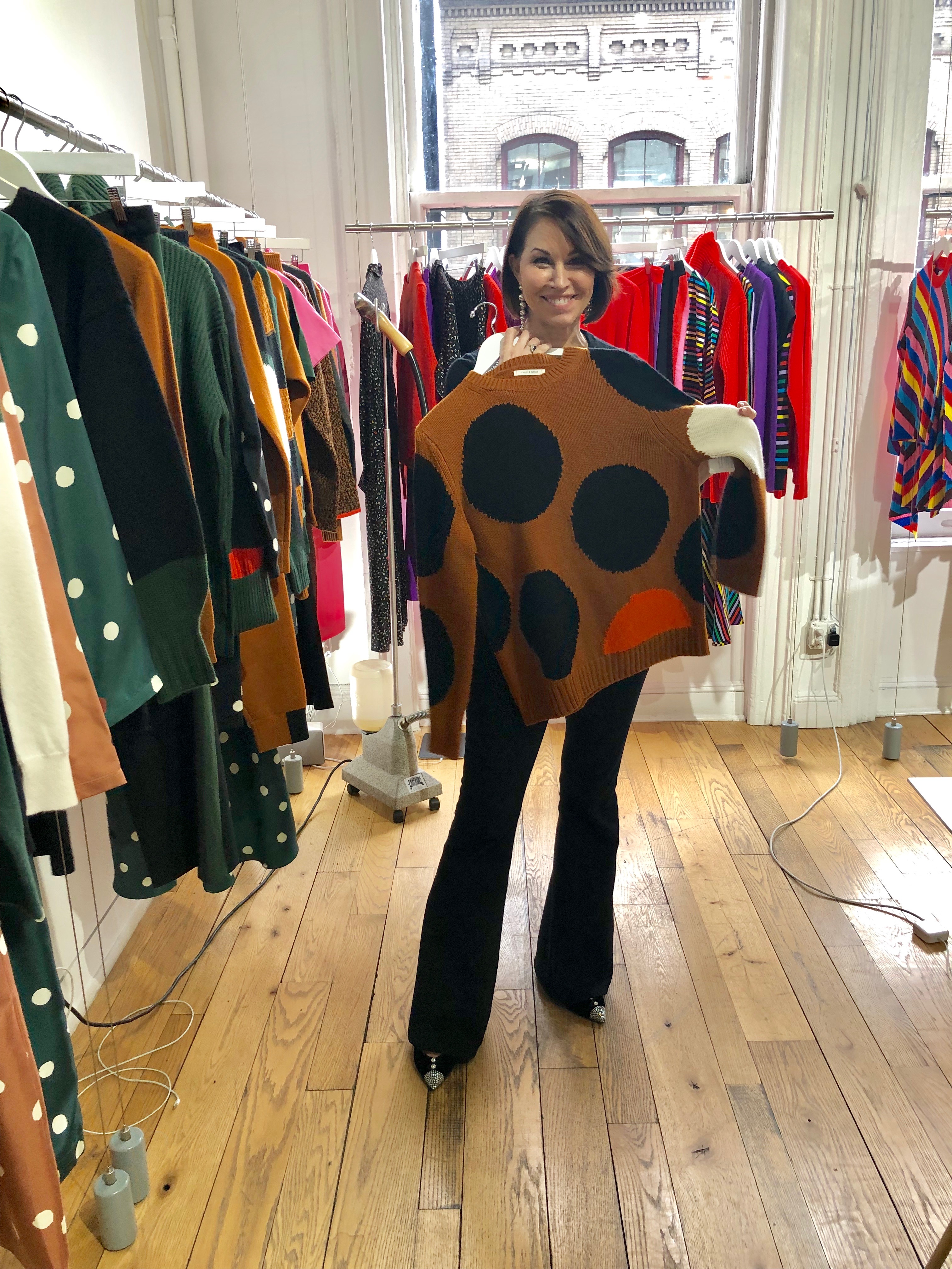 Woman with brown hair holding up a polka dot camel sweater with black and red polka dots in a showroom in Manhattan