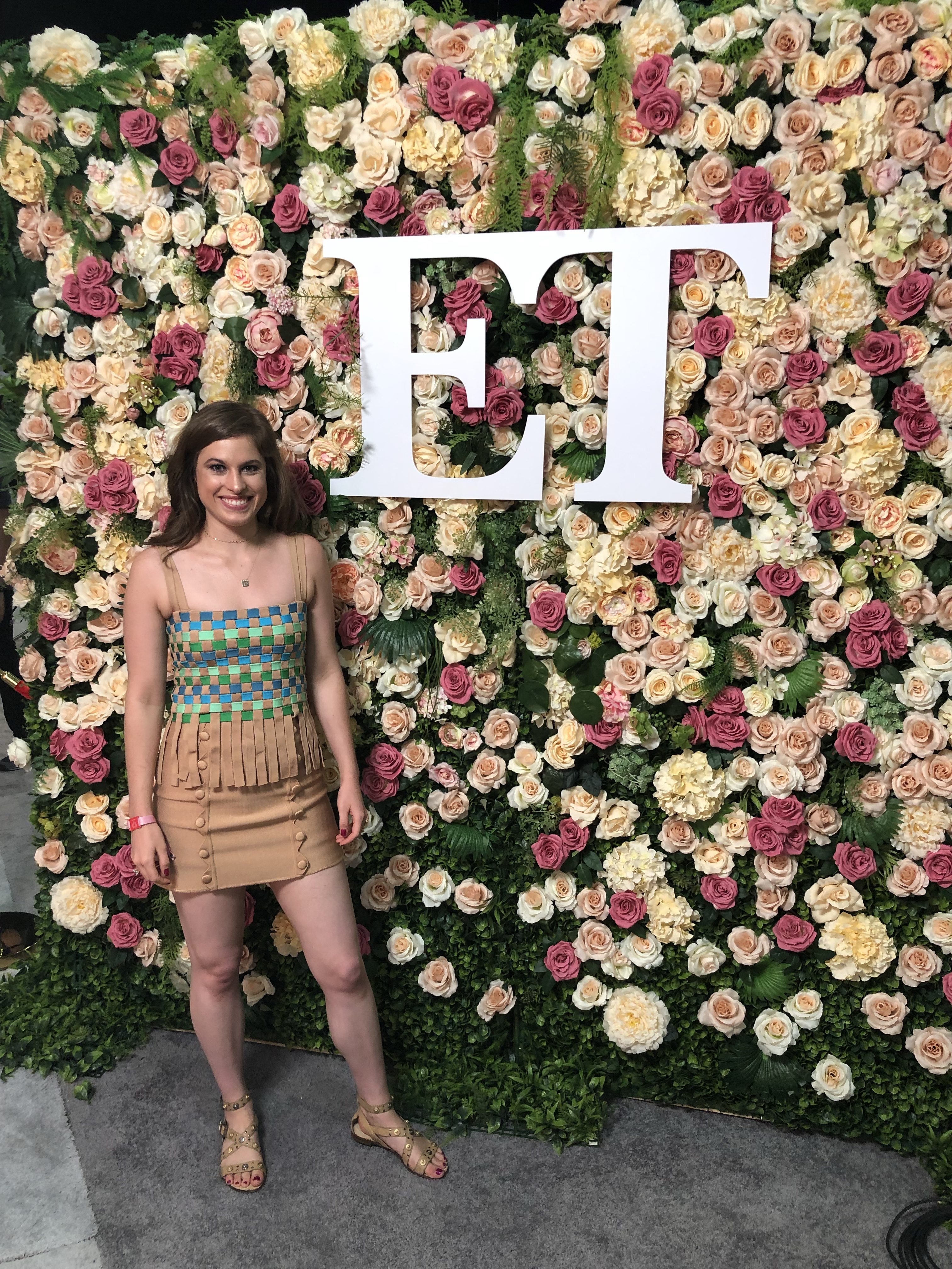 Woman standing in front of a flower backdrop wearing a printed spaghetti strap top and a beige mini skirt and sandals. She has brown curly hair
