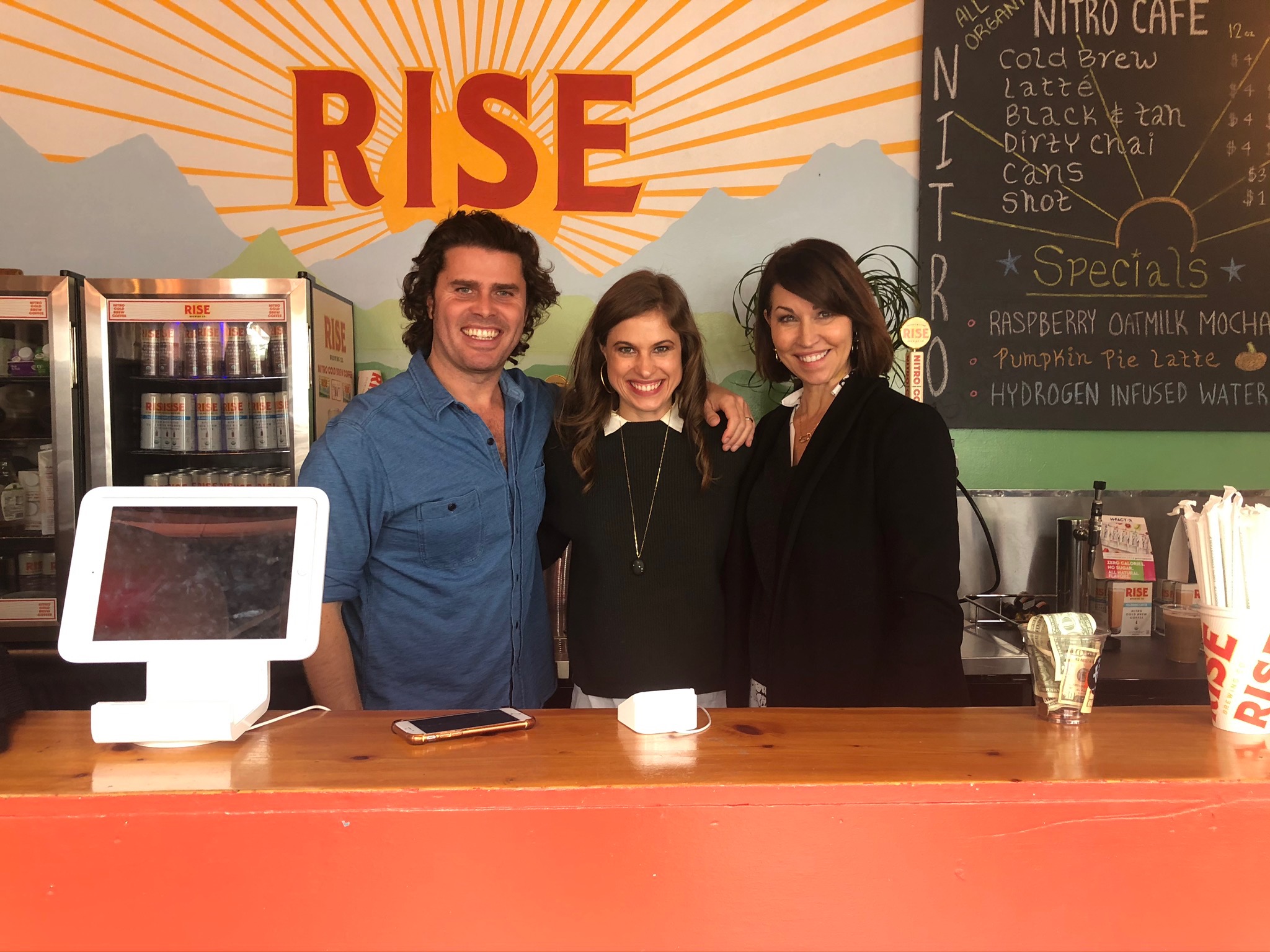 Three people at the RISE cafe in Lower East Side Manhattan: a man with a denim button down shirt, a woman with a dark green sweater and a woman with a black blazer on: all with brown hair