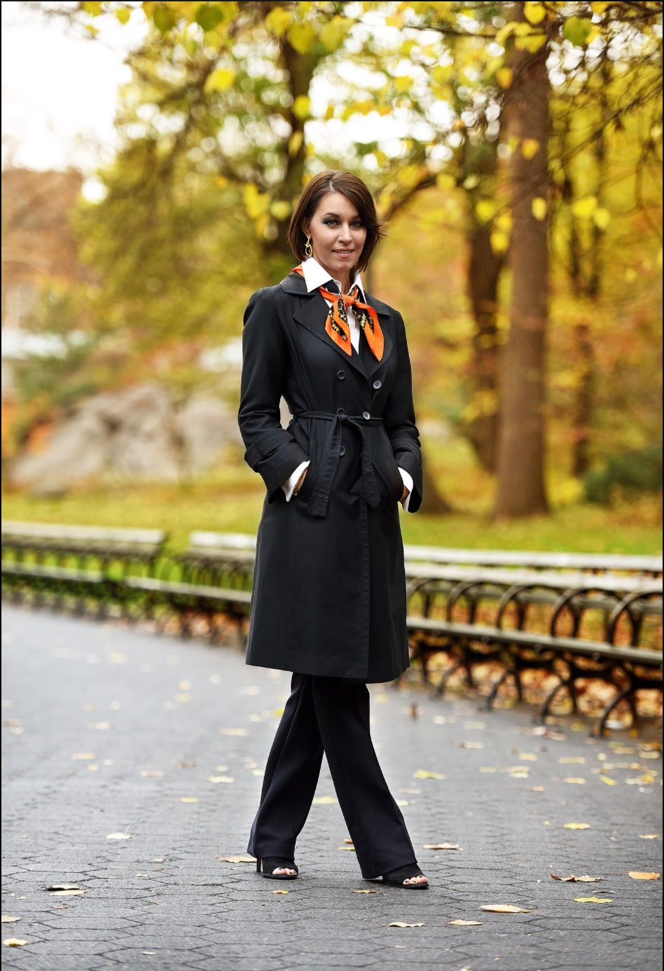 Woman wearing a black trench coat and an orange hermes scarf walking in central park in the fall