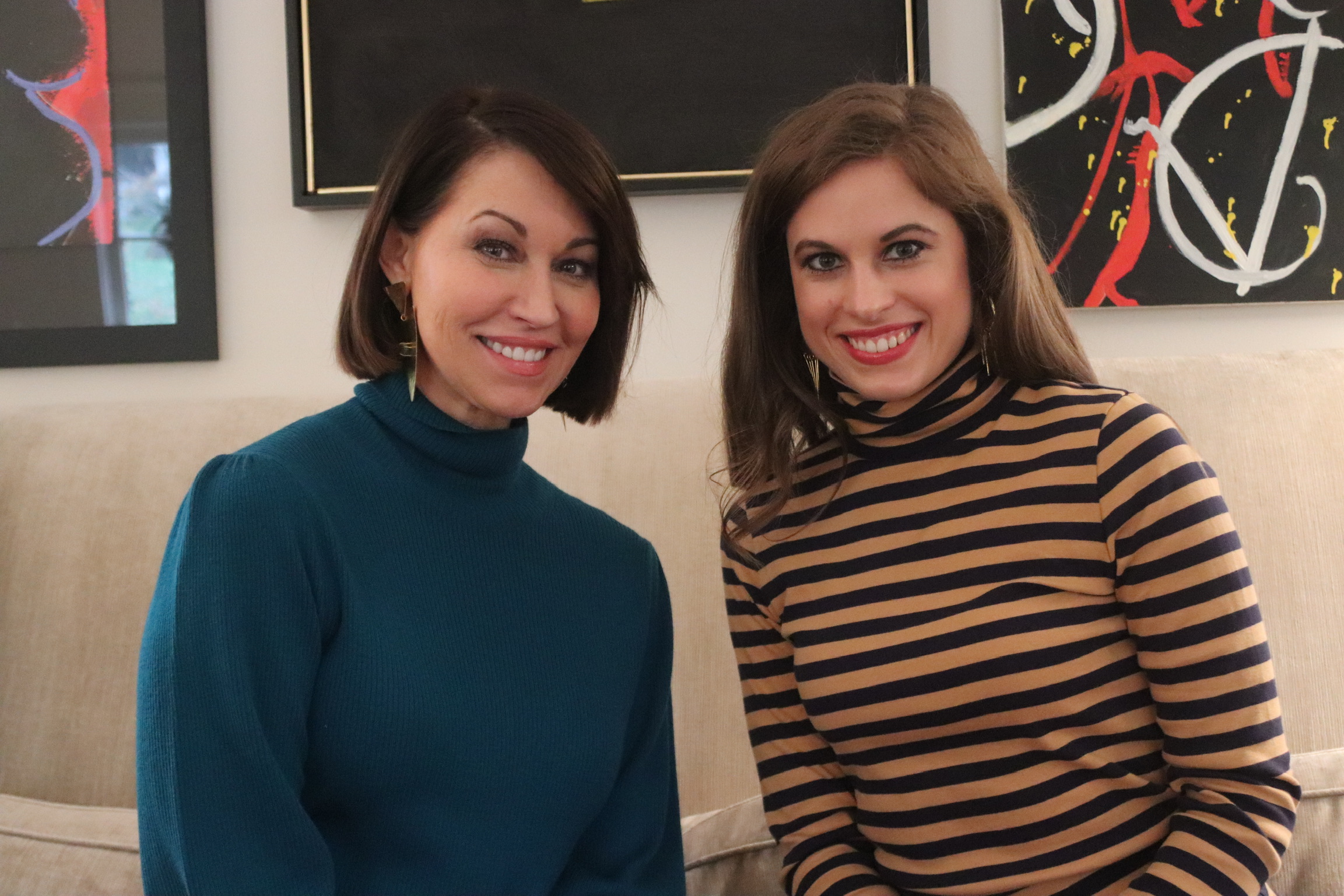 Two women sitting on a sofa: they both have brown hair. One is wearing a teal turtleneck sweater and the other is wearing a black & gold stripe turtleneck