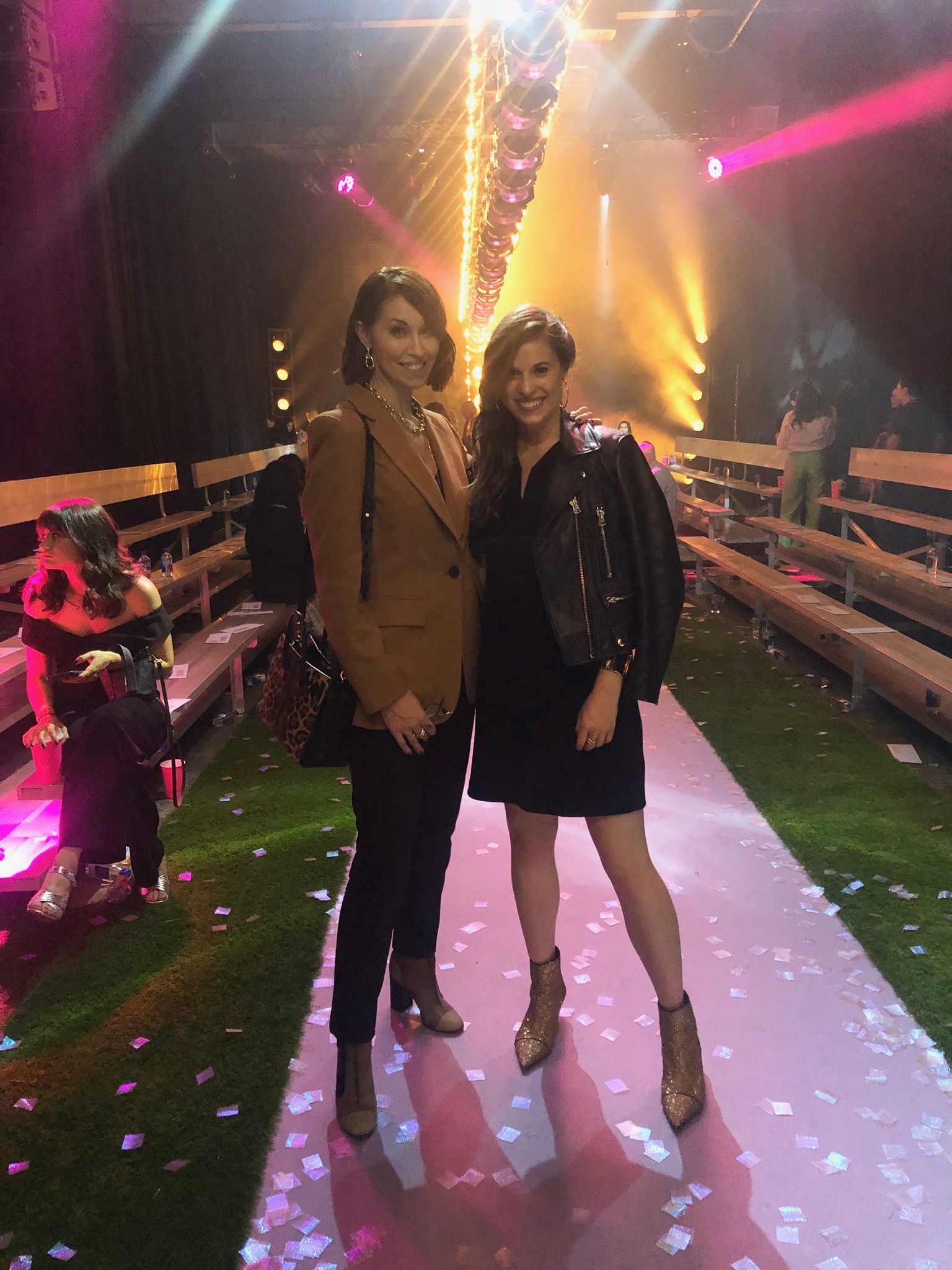 Two women at a fashion show. One wearing a camel blazer & black jeans and booties. The other wears a black bomber & black dress with booties.
